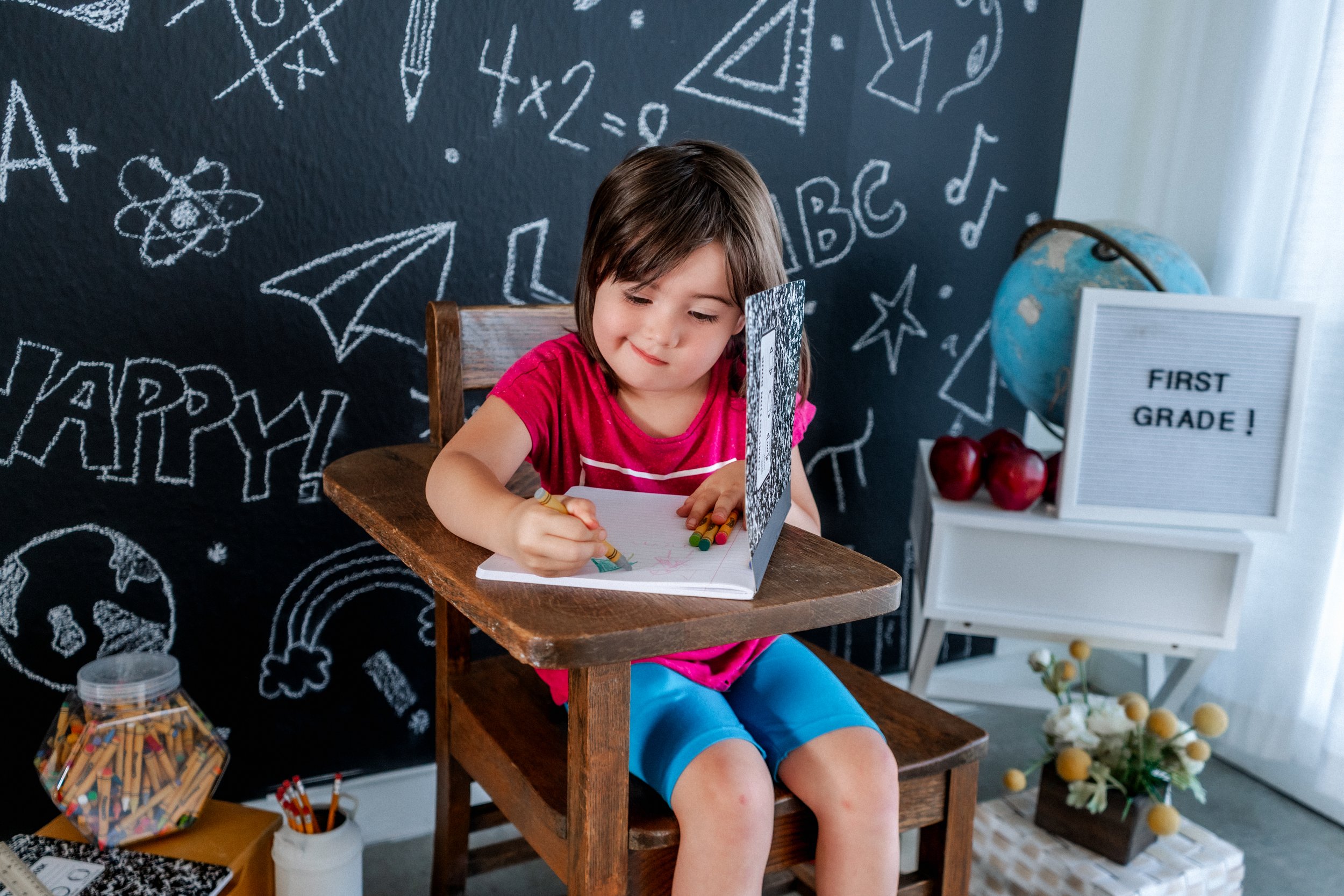 Young girl sitting at a wooden desk drawing with colored pencils, in front of a chalkboard with educational doodles. "First Grade!" sign and globe in the background.