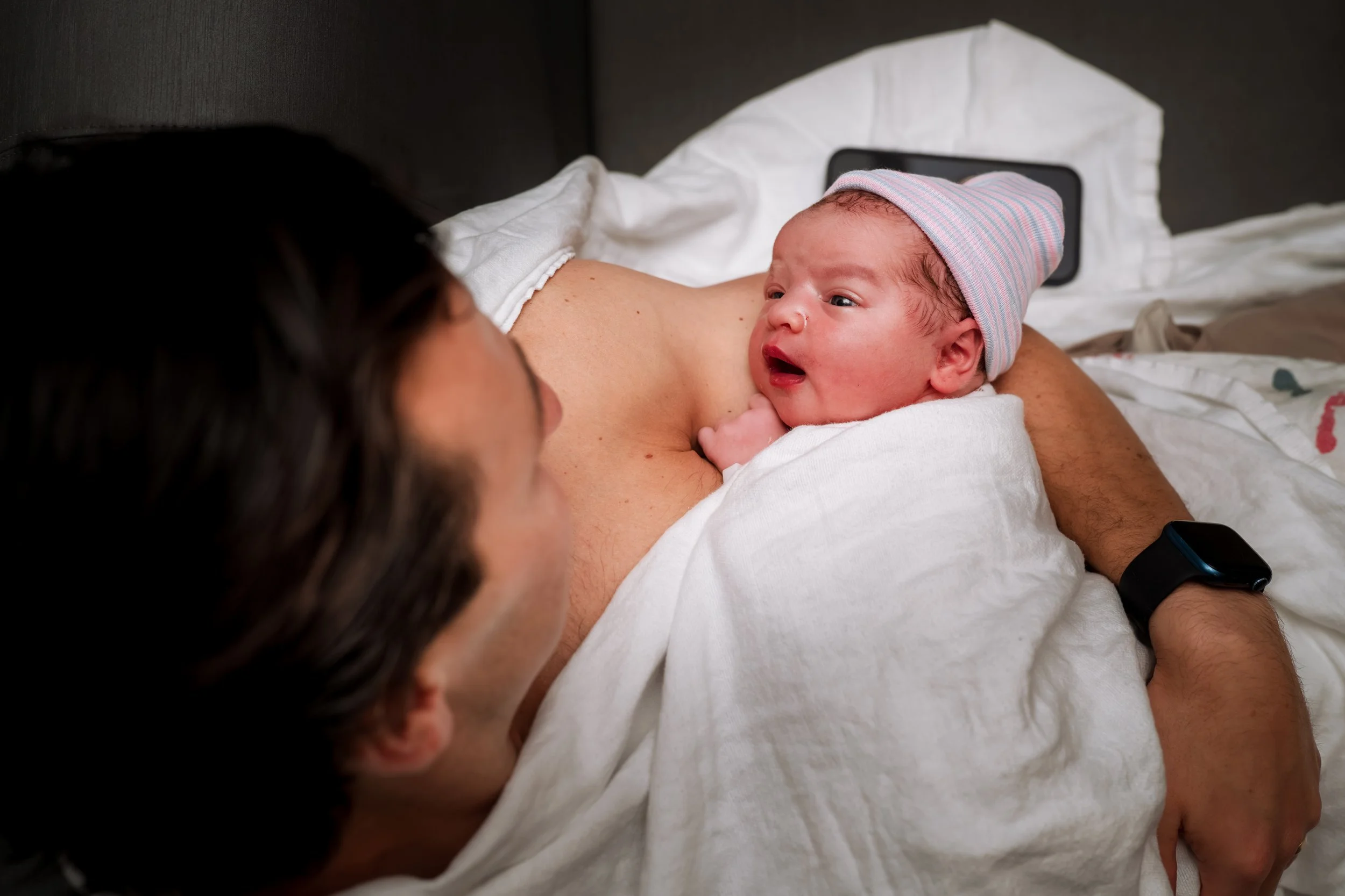 A newborn baby with a pink and blue striped hat lying on an adult's chest, looking at the adult with a curious expression, while the adult lies on a bed and wears a white shirt and a black smartwatch.