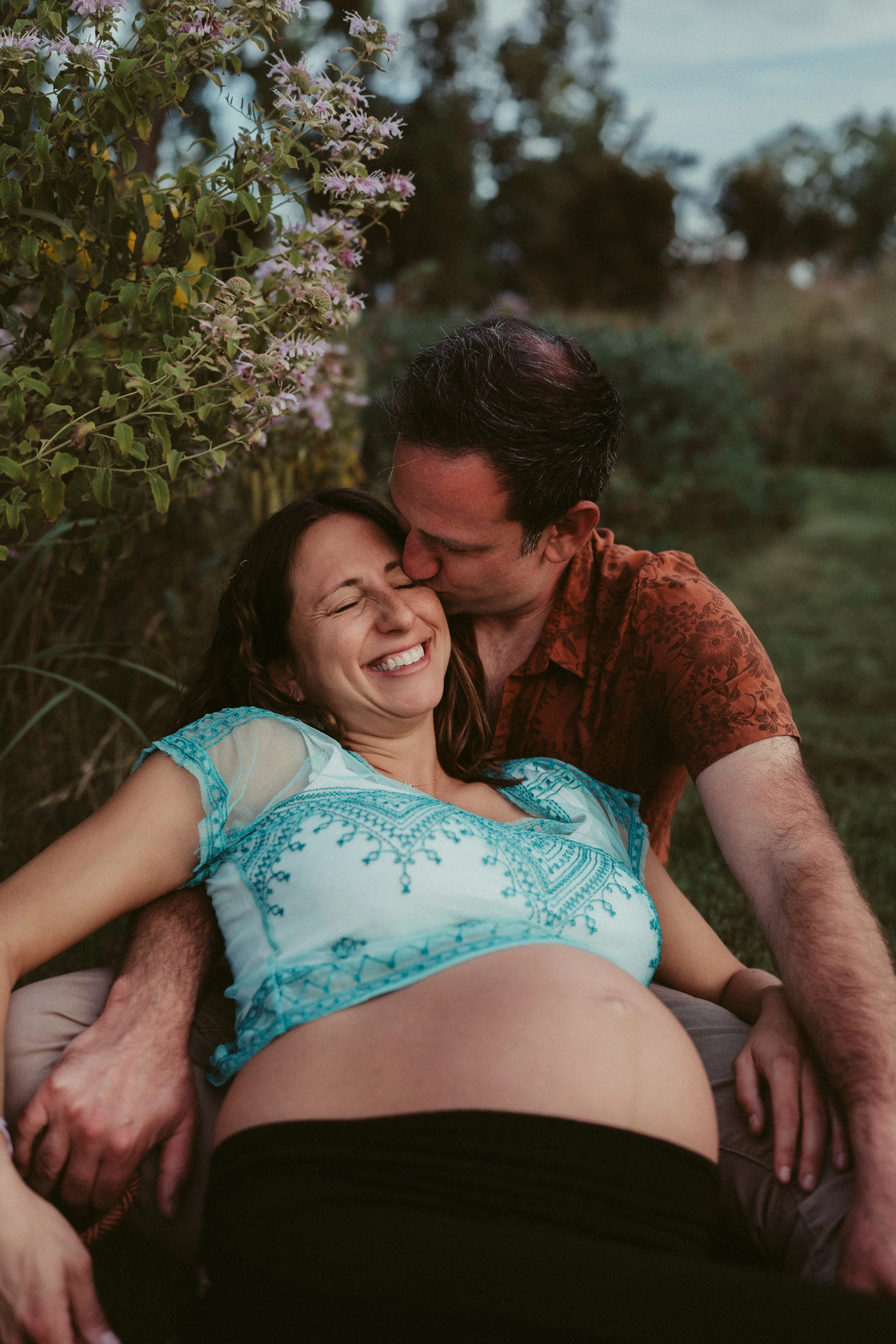 A joyful woman with dark hair and a nose piercing smiling with her eyes closed, sitting outside, with a man kissing her on the forehead. The woman is wearing a light blue and white embroidered top, and the man has dark hair and is wearing a patterned