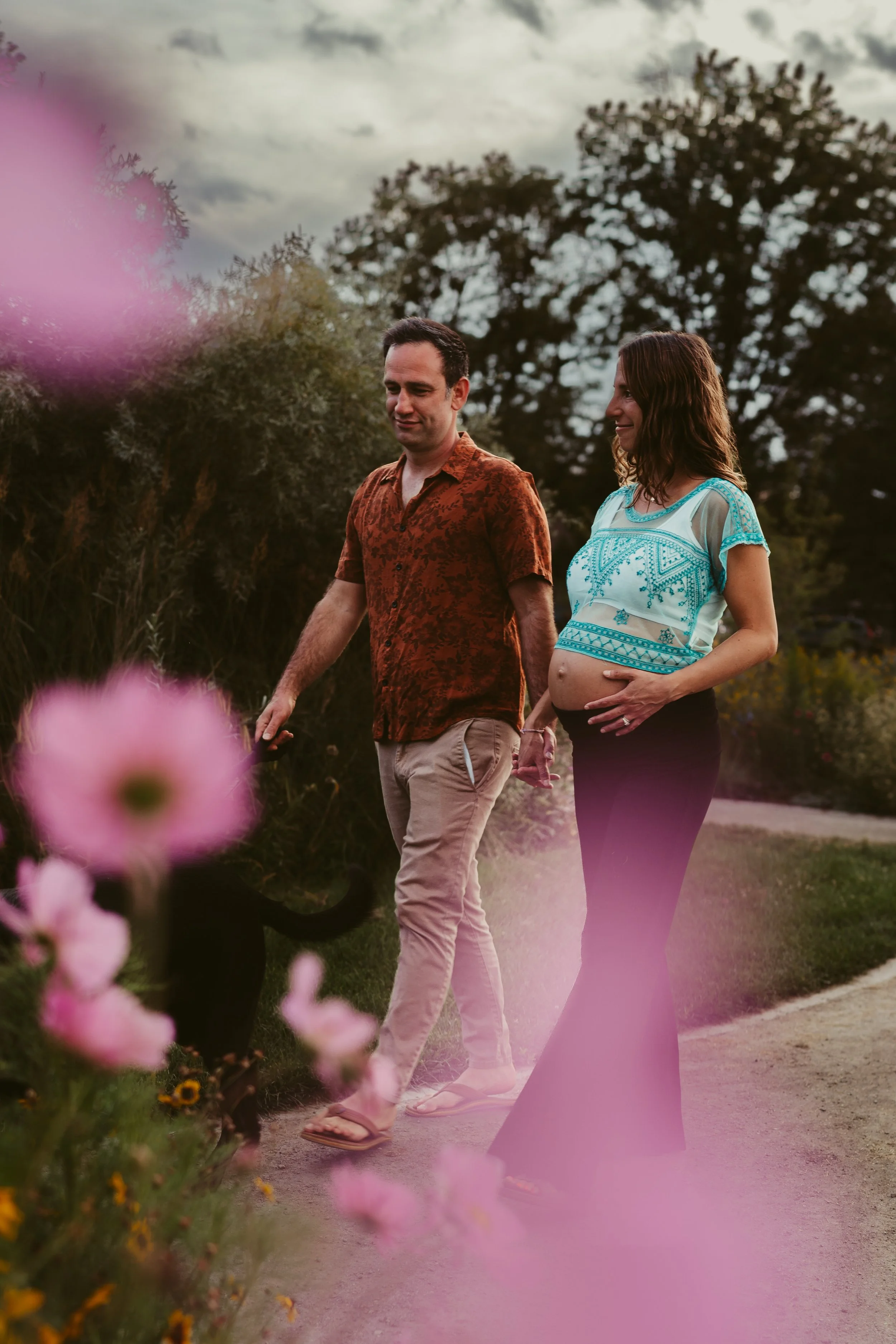 A pregnant woman and a man walking outside on a pathway, with flowers in the foreground and trees in the background during cloudy weather.