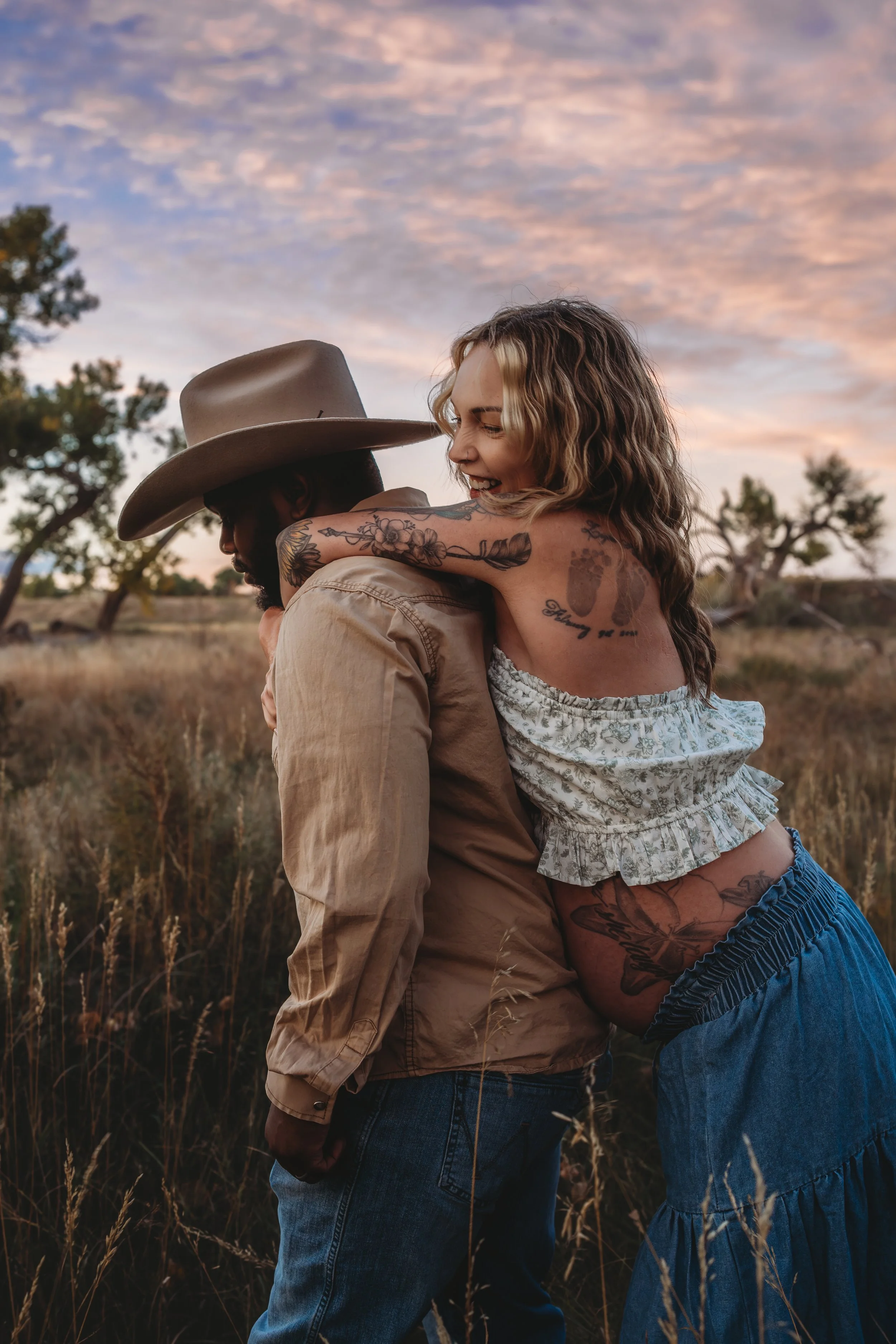 A couple embracing outdoors at sunset, with the woman smiling and looking happy, and the man wearing a cowboy hat, in a field with trees and a colorful sky in the background.