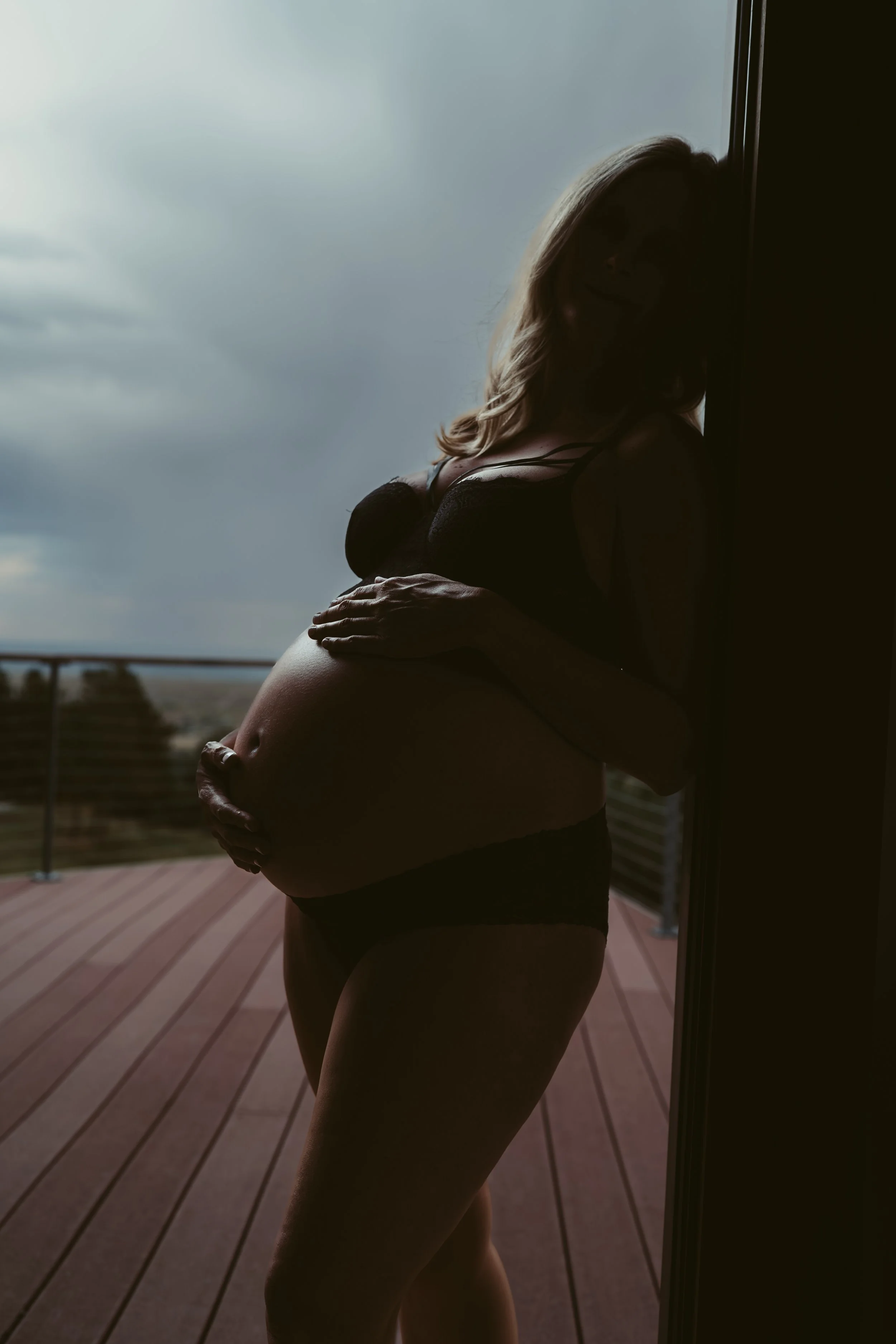 Silhouette of a pregnant woman holding her belly, standing on a wooden deck with cloudy sky in the background.