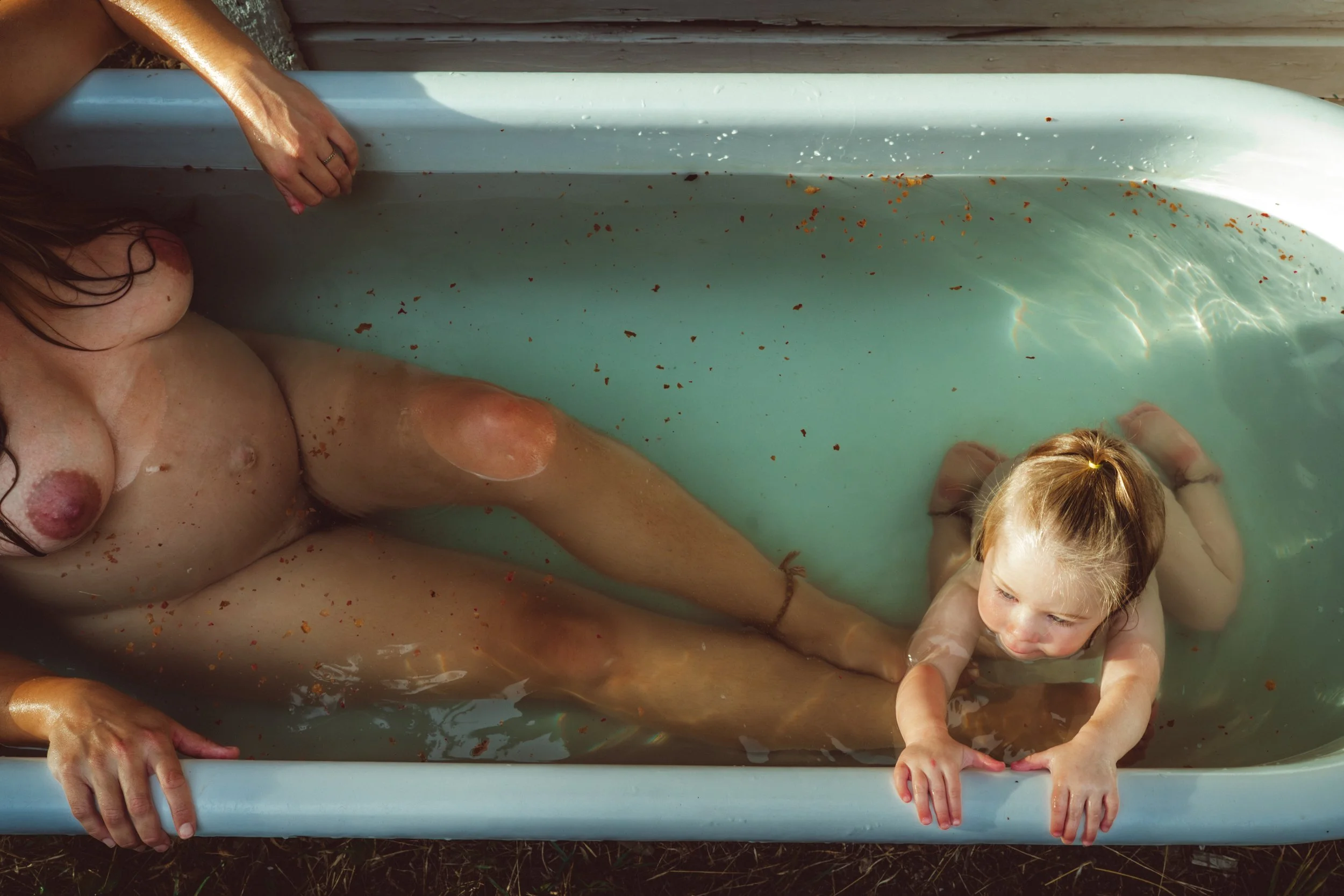 A woman and a child are taking a bath together in a small plastic tub filled with water. The woman is on the left side, and the child is on the right side. Both are holding the edge of the tub with their hands, and some water and dirt particles are v
