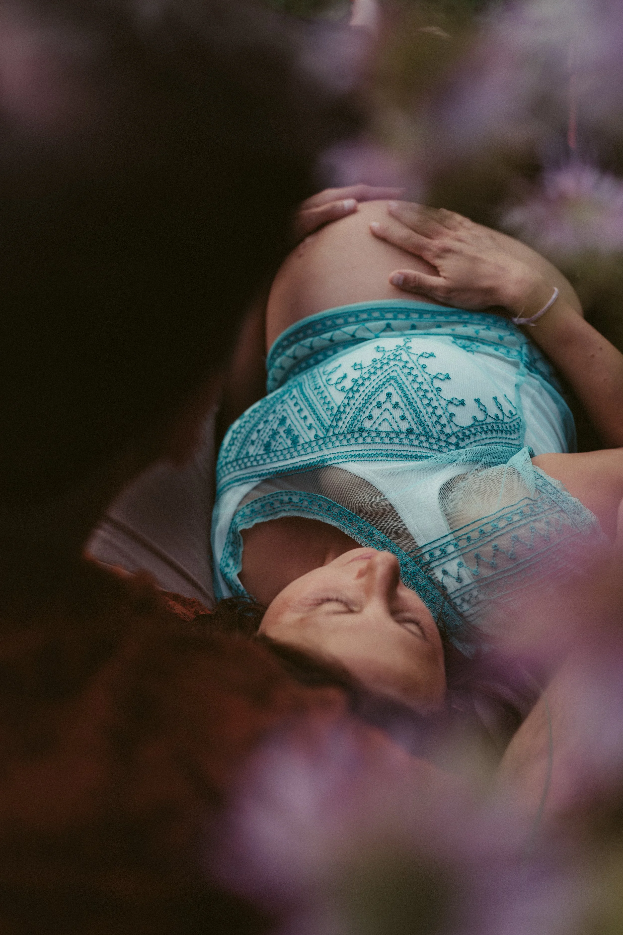 A woman lying on a bed with her hand on her pregnant belly, wearing a white and blue embroidered dress, with blurry pink and purple flowers in the foreground.