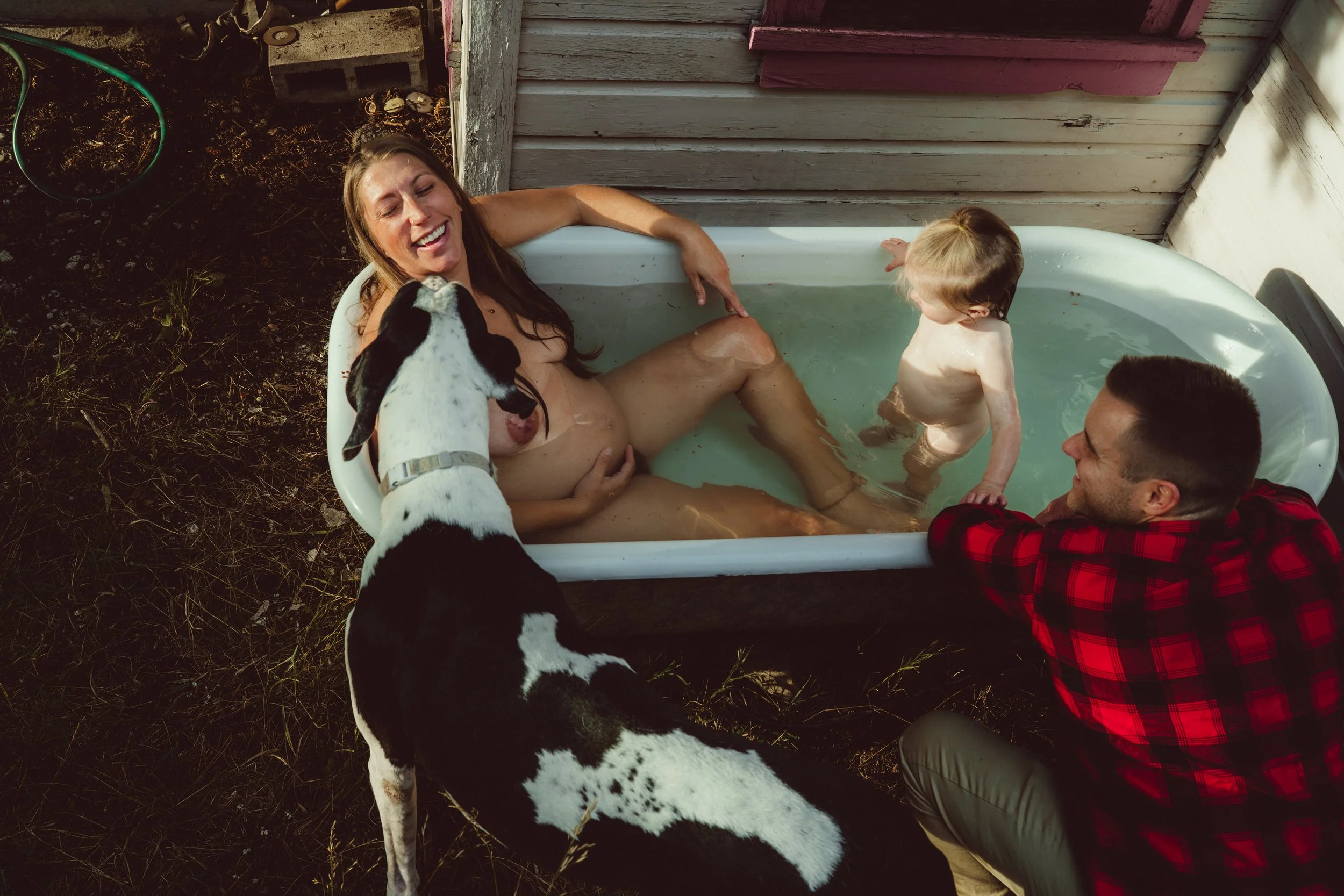 A woman and a man relax in a bathtub outside, laughing, with a young child standing in the water. A black and white dog is leaning over the woman's lap.