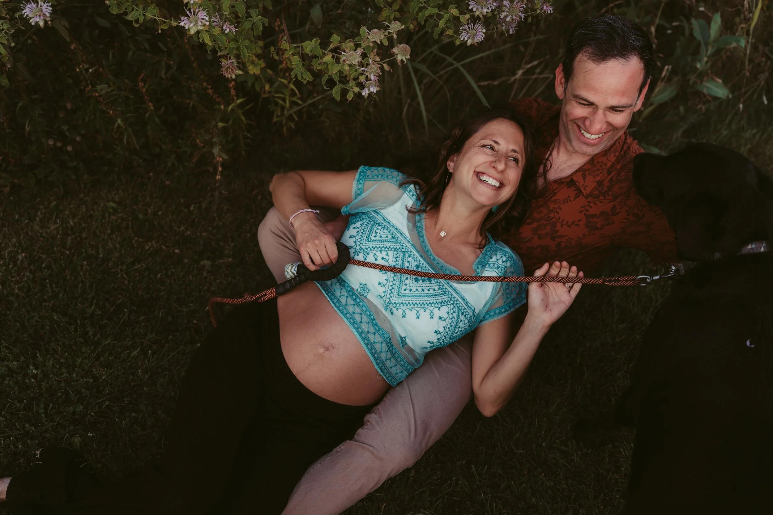 Smiling woman lying on her back outdoors with a pregnant belly, next to a man, both happy, in a garden area with purple flowers and green foliage.