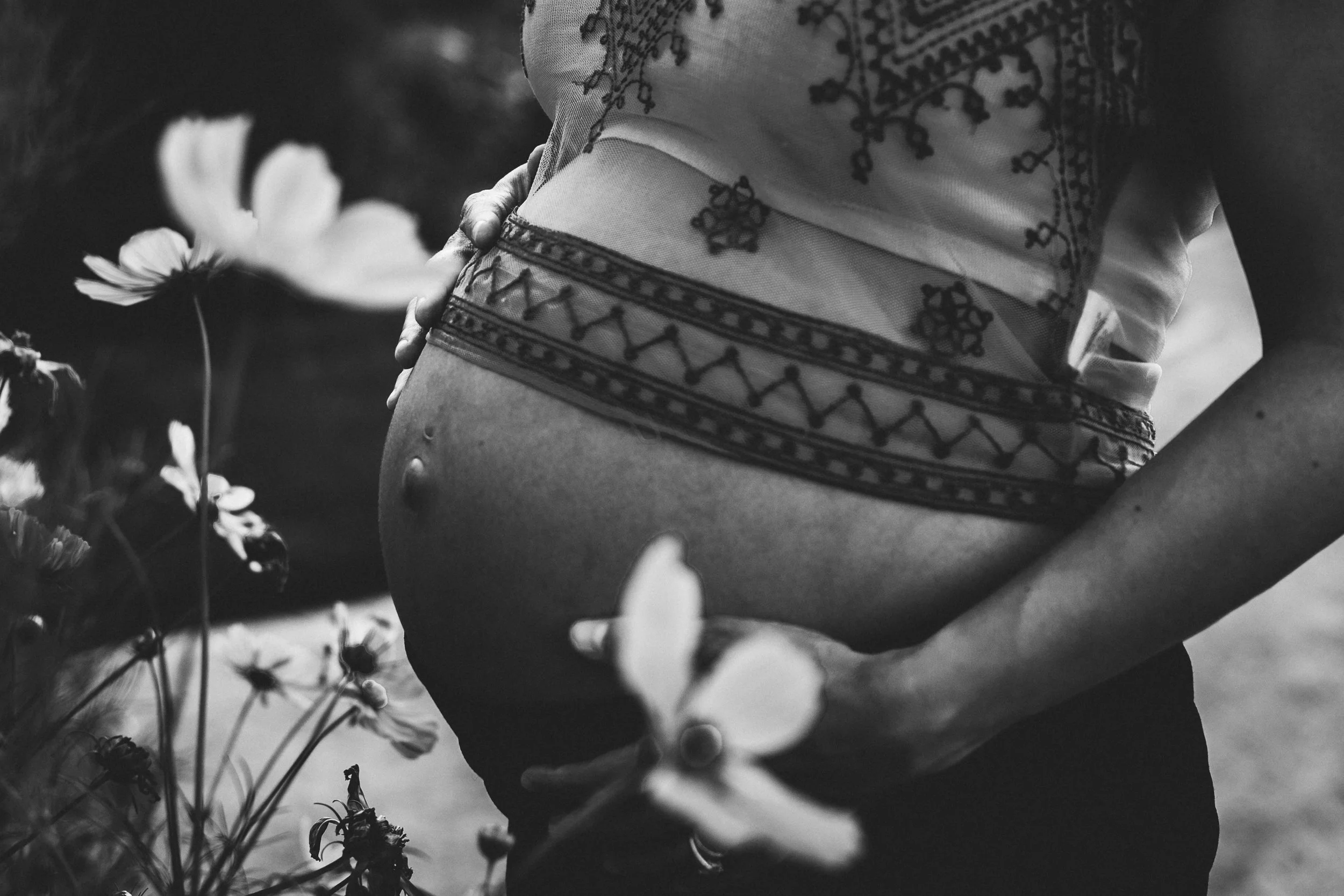 Black-and-white close-up photo of a person wearing embroidered shorts, with hand resting on their hip, surrounded by flowers.