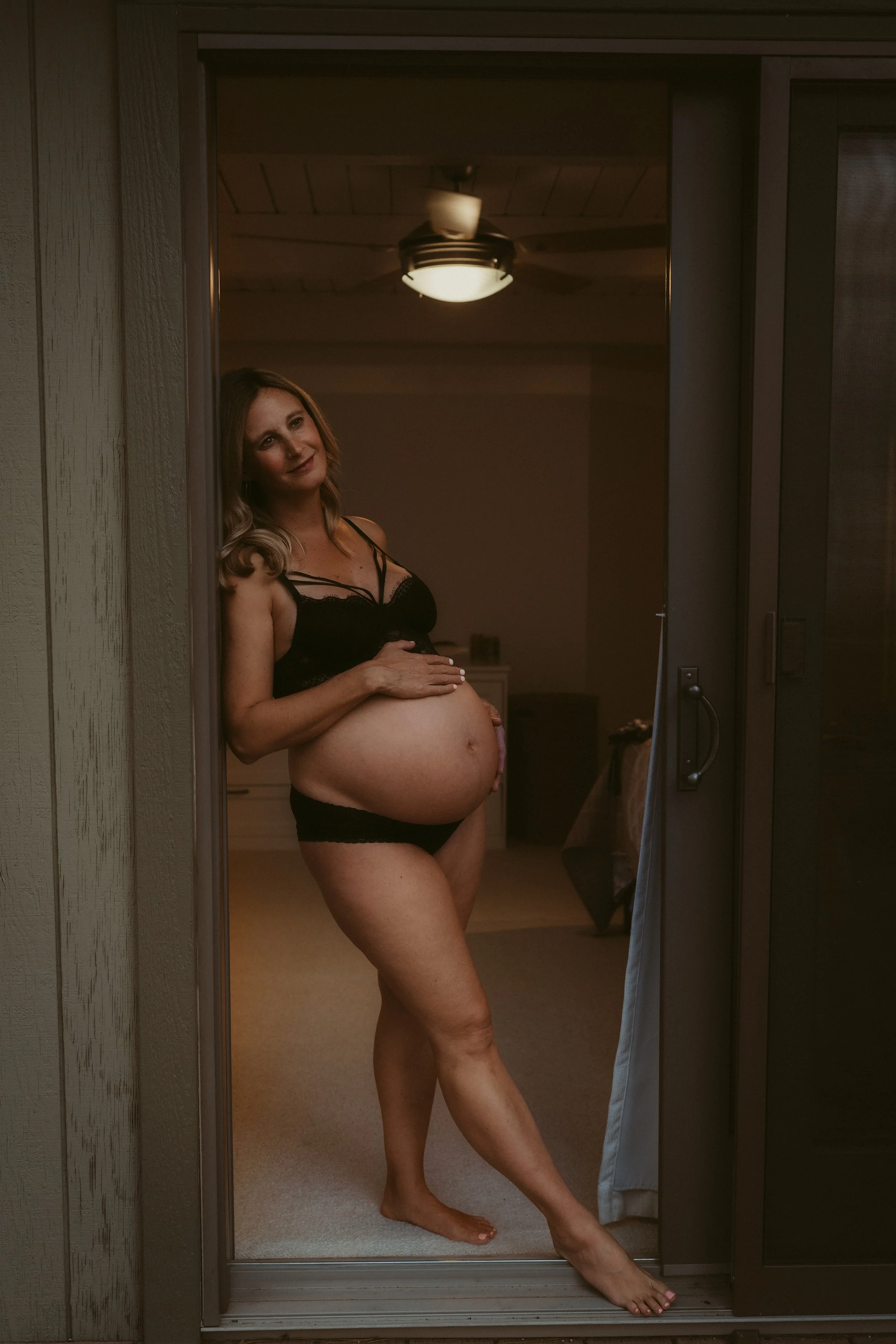 Pregnant woman wearing black lingerie standing in a doorway, looking outside.