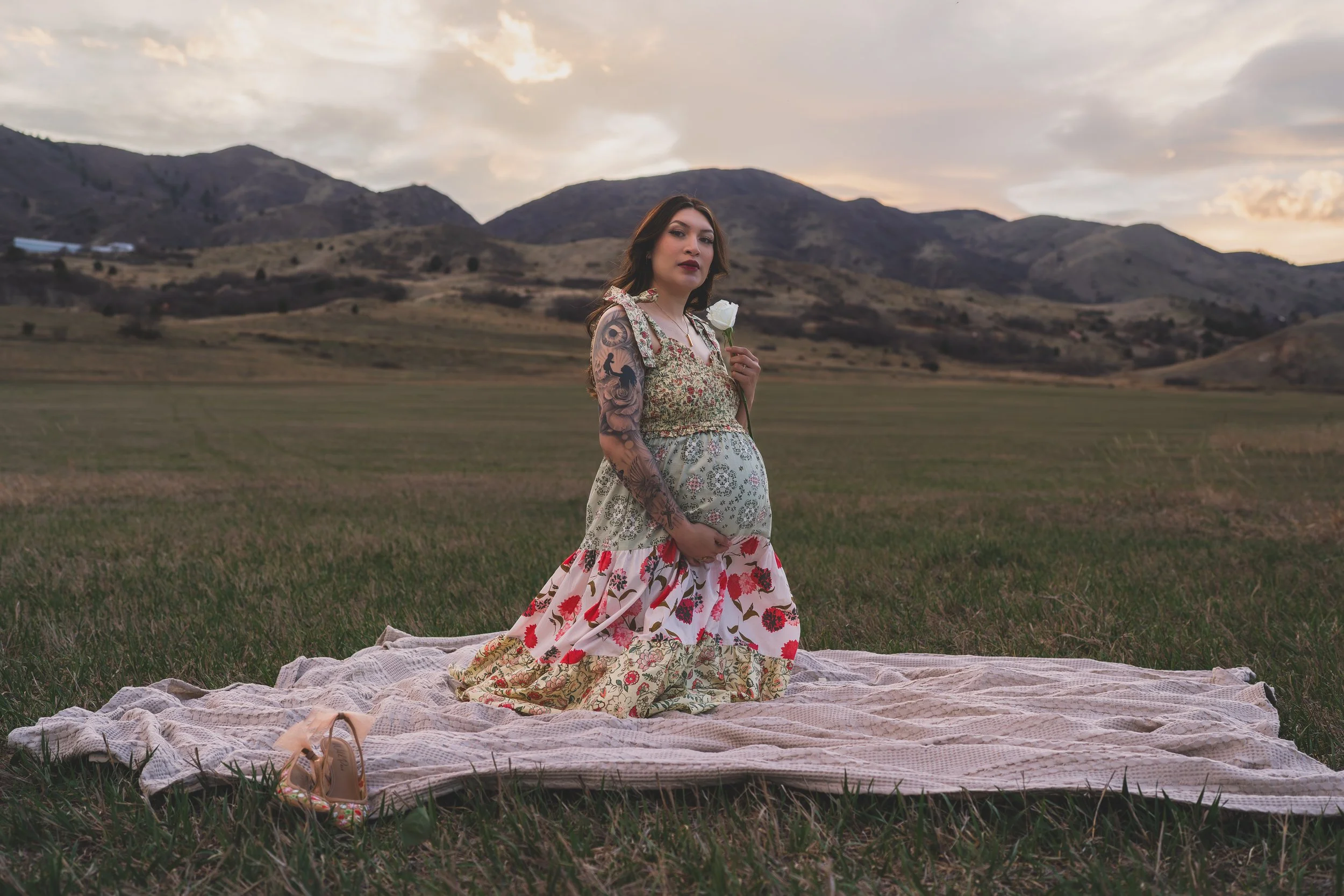 A woman with tattoos in a floral dress holding a white rose stands on a blanket in a grassy field with mountains and a cloudy sky in the background during sunset.