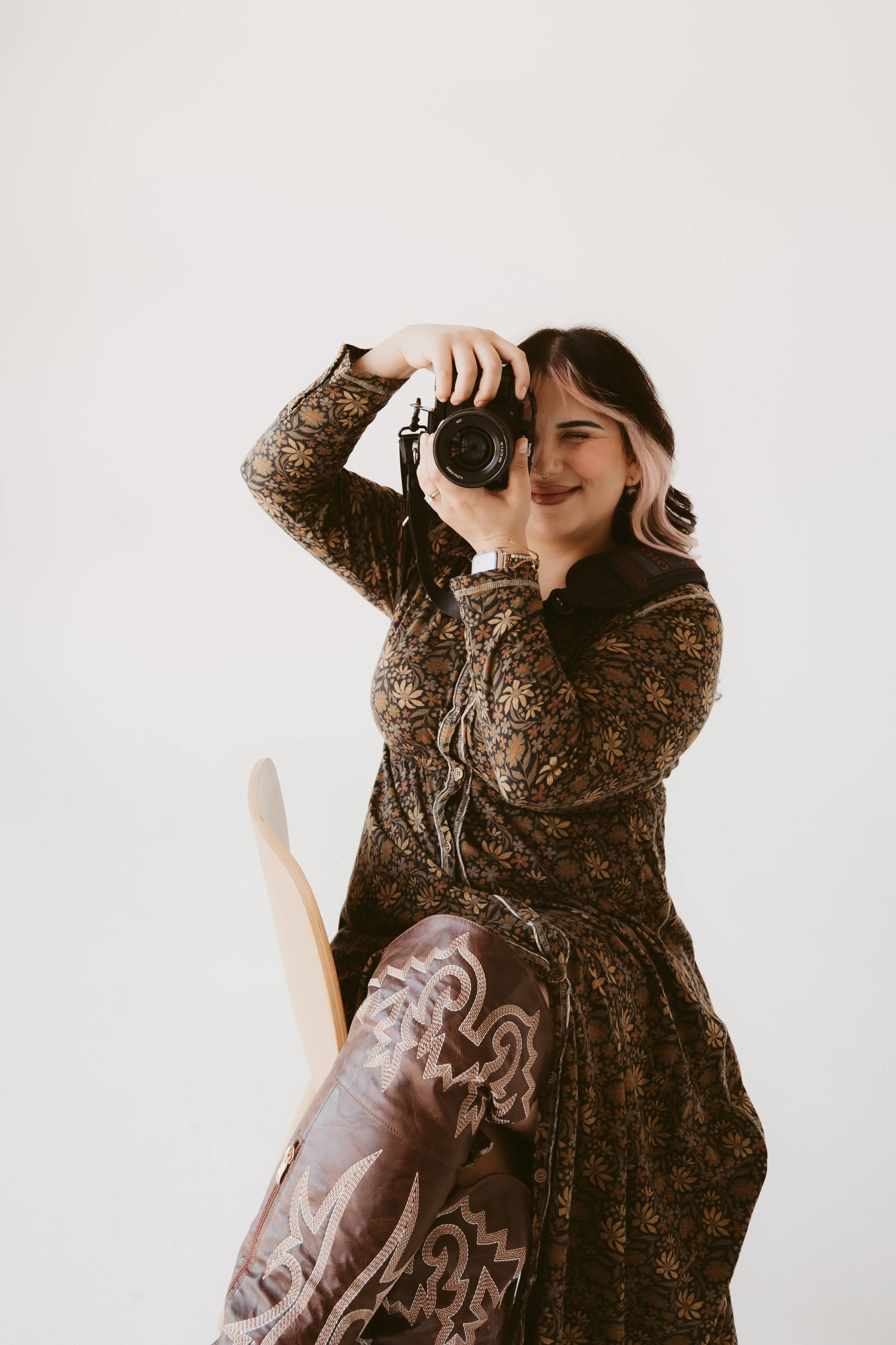 A young woman with pink and black hair taking a selfie with a camera, smiling with her eyes closed, wearing a patterned long-sleeve dress, sitting on a chair against a plain white background.