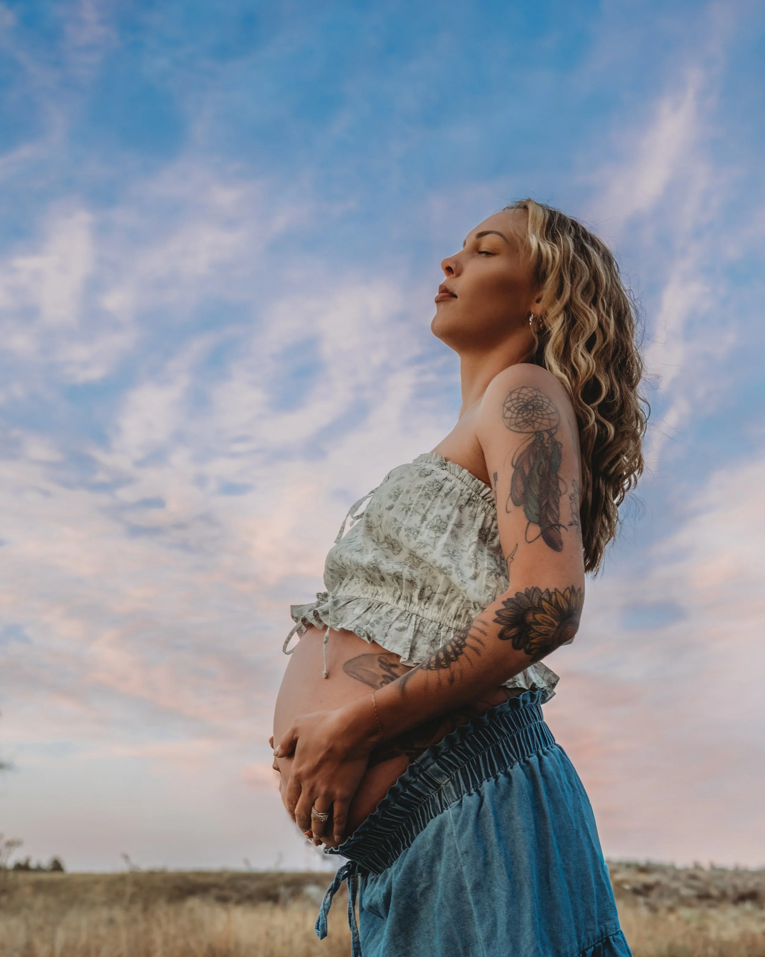 A woman with curly blonde hair and tattoos on her arm and shoulder stands outdoors with her eyes closed, facing the sky at sunset or sunrise. She is wearing a strapless top and blue shorts, and the background features a grassy field and a sky with cl