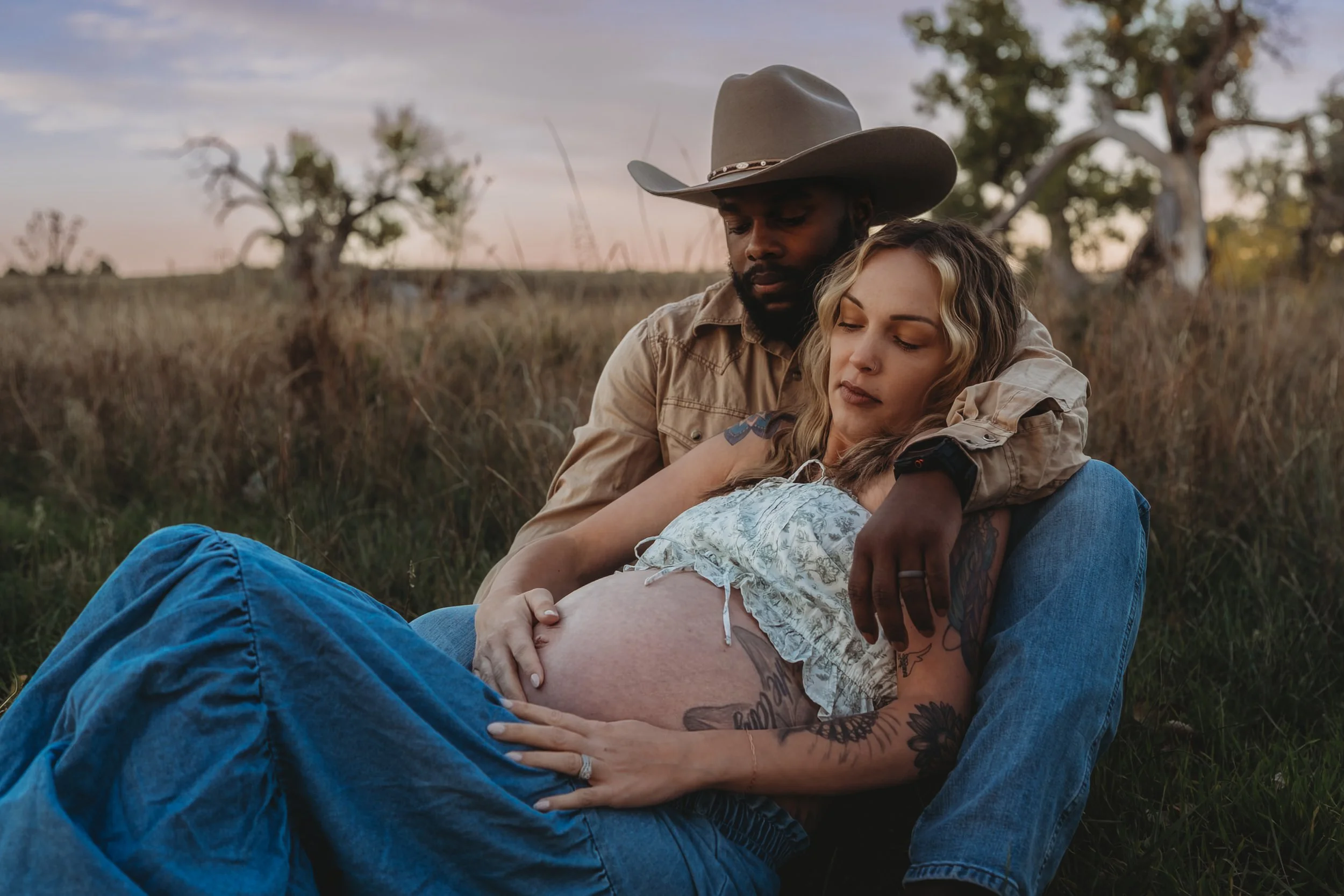 A couple sitting in a field during sunset, with the man wearing a cowboy hat and the woman resting her head on his chest, both appearing relaxed and content.