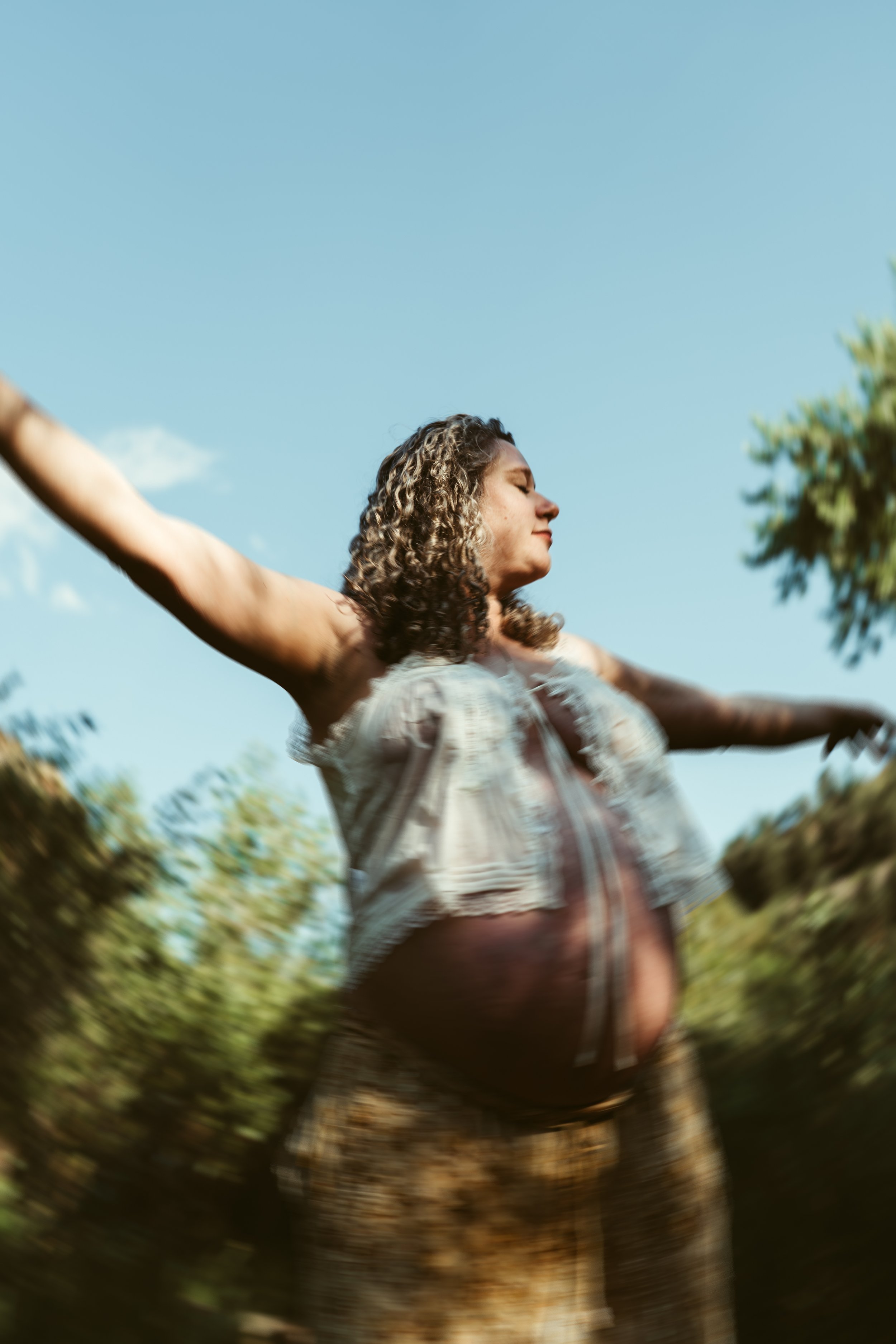 Pregnant woman with curly hair wearing a light-colored top and patterned skirt standing outdoors with arms outstretched and eyes closed against a blue sky.