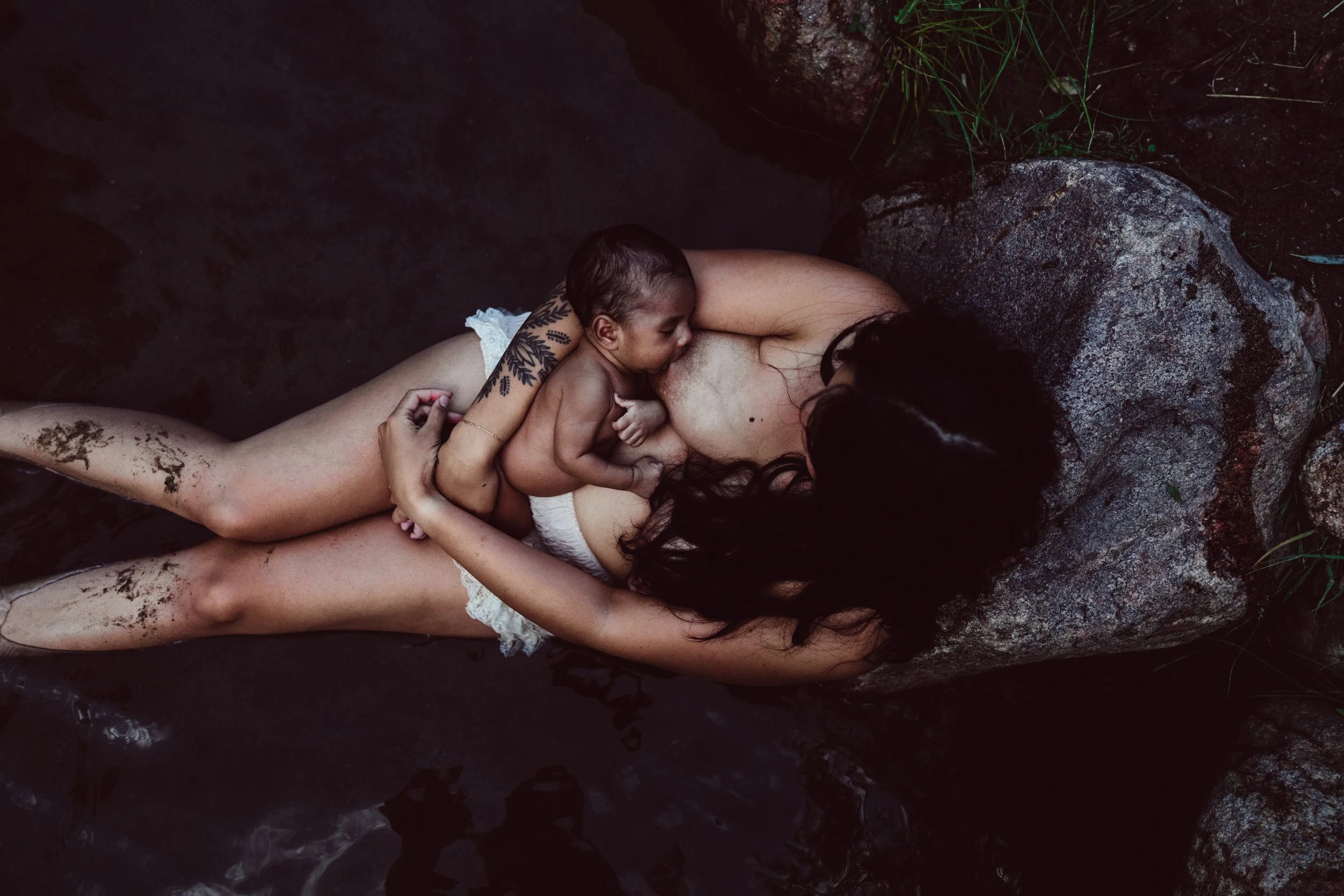 A woman with dark hair and tattoos lying in a water body with a small child breastfeeding, surrounded by rocks and green plants.