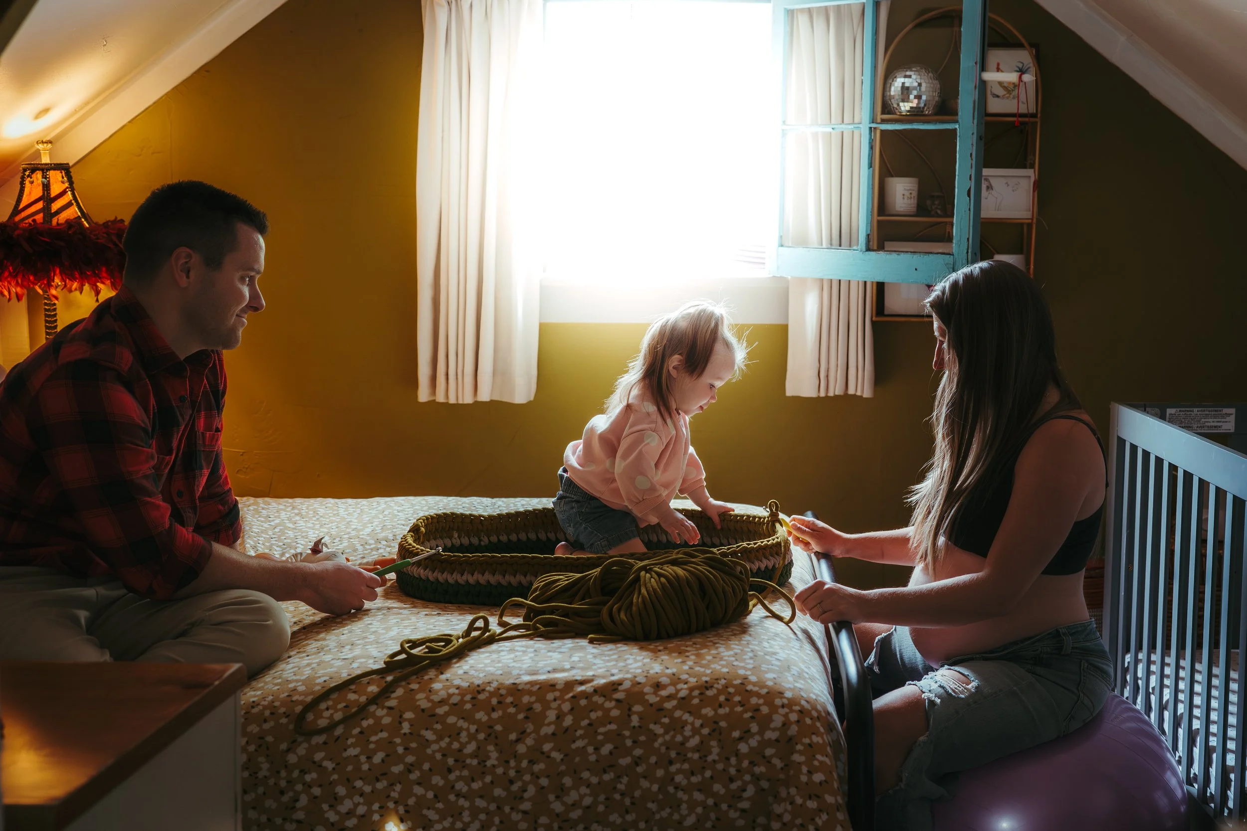 A family of three with a man, a woman, and a young girl in a bedroom, knitting together with yarn and knitting needles.