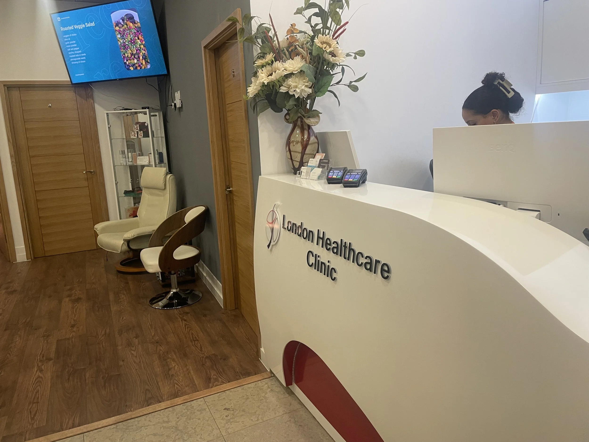 Reception area of London Healthcare Clinic with a curved white reception desk, a large floral arrangement, two chairs, and a woman working on a computer behind the desk. A wall-mounted screen displays a menu for roasted veggie salad.