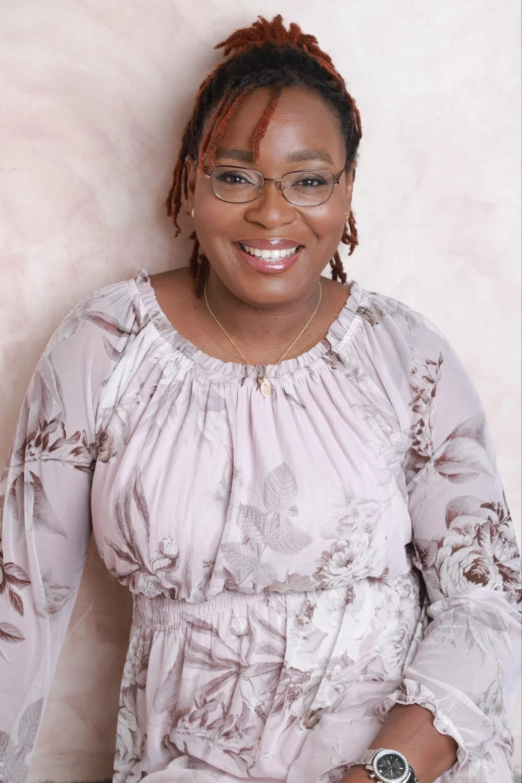 A smiling woman with glasses, wearing a light floral blouse and a watch, standing against a light-colored wall.