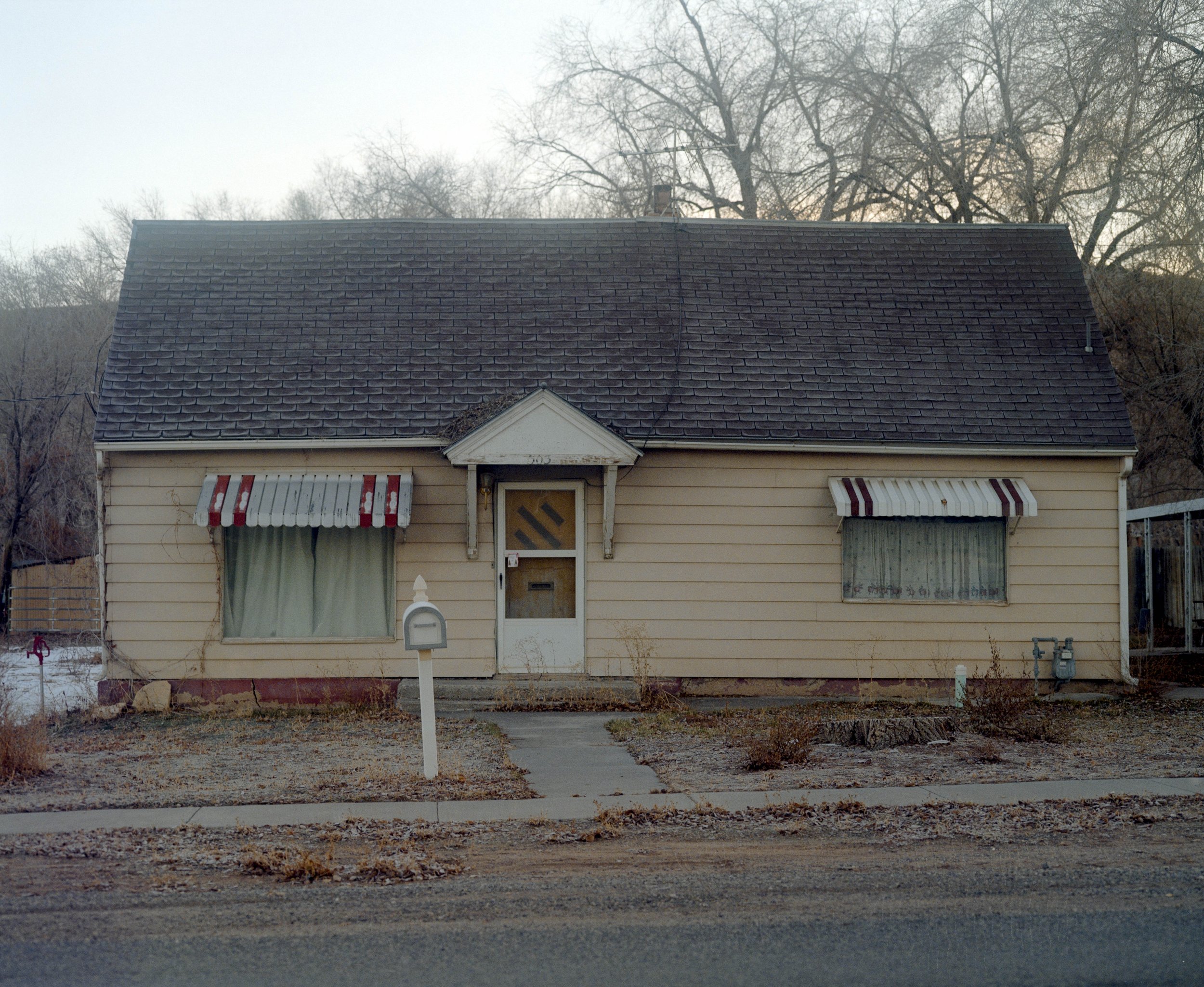 Une maison simple avec un toit en tuiles noires, des volets à rayures rouges et blanches, et des rideaux verts derrière les fenêtres. La maison a une petite entrée avec un auvent blanc et un jardin peu entretenu devant.