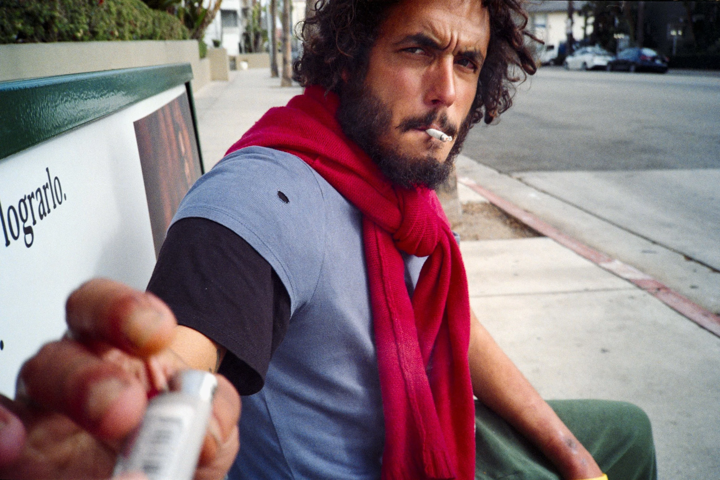 Homme barbu avec cheveux bouclés, portant un t-shirt gris, une veste noire, une écharpe rouge, assis sur un banc de parc, tenant un marqueur, cigarette dans la bouche, regardant la caméra.