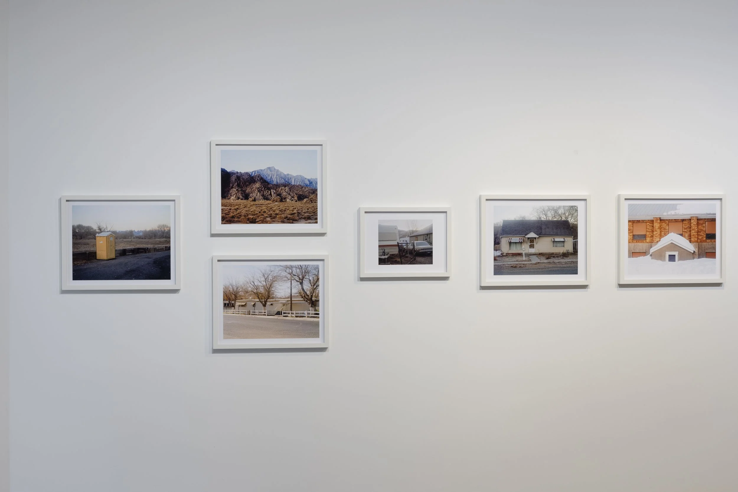 Six photographies de paysages et de scènes de vie urbaine exposées sur un mur blanc d'une galerie d'art.