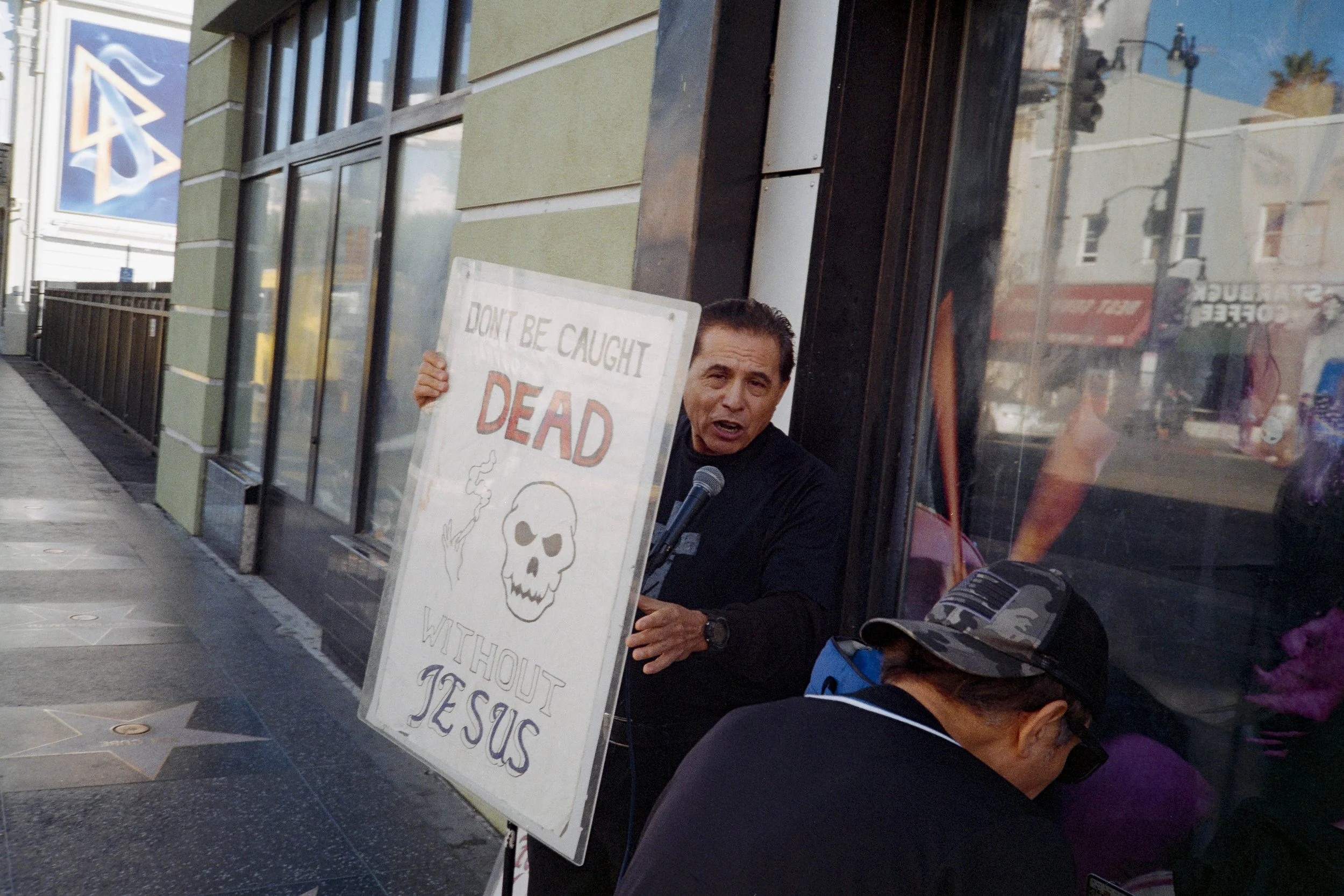 Un homme parle en public devant un panneau qui dit "Ne sois pas pris pour mort sans Jésus" en anglais, avec une illustration d'un crâne. La scène se déroule dehors le long d'un trottoir.