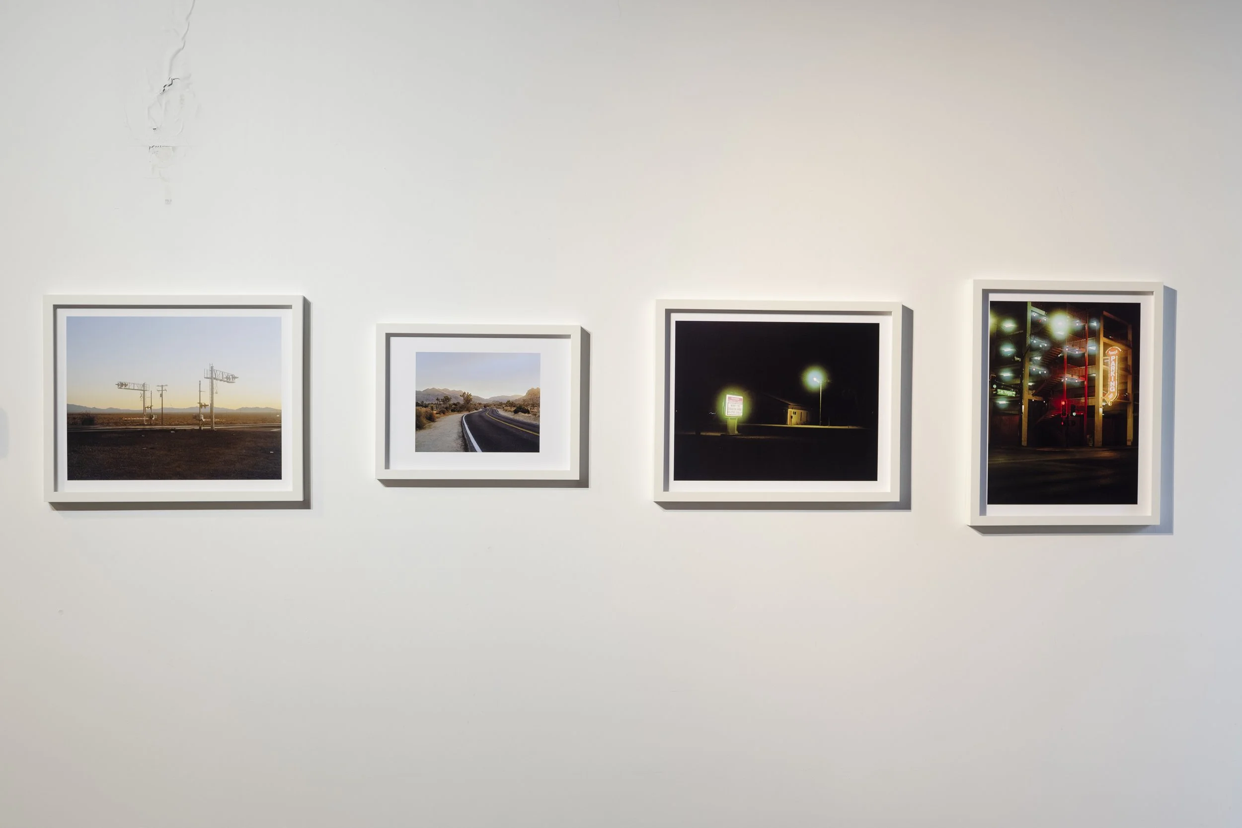 Quatre photographies encadrées exposées sur un mur blanc, représentant des scènes nocturnes et diurnes avec des paysages et des lumières urbaines.
