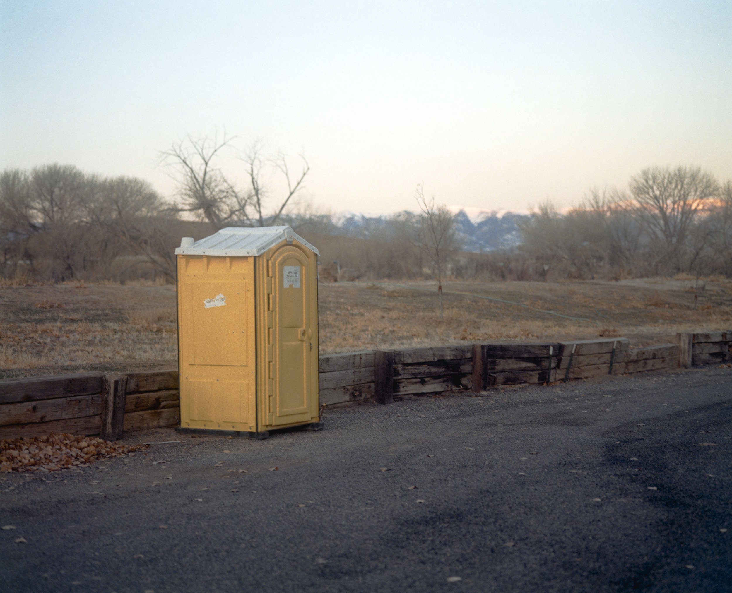 Une cabine de toilette jaune installée sur le bord d'une route, avec un paysage de arbres dénudés en arrière-plan et des montagnes au loin.