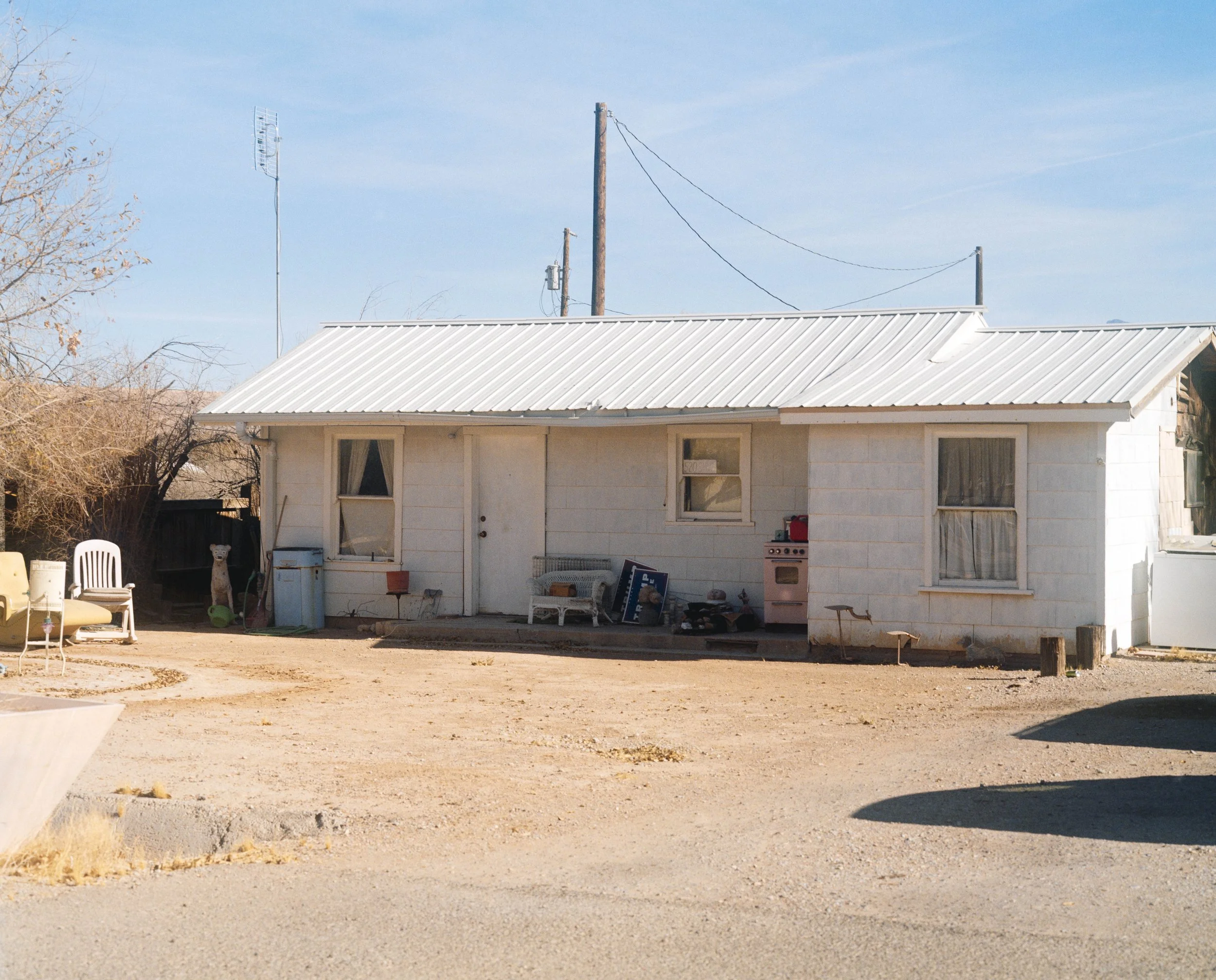 Maison simple en pierre avec une toiture en métal blanc, fenêtres à rideaux, devant un sol de terre, avec des meubles et des jouets à l'extérieur, et un ciel clair.