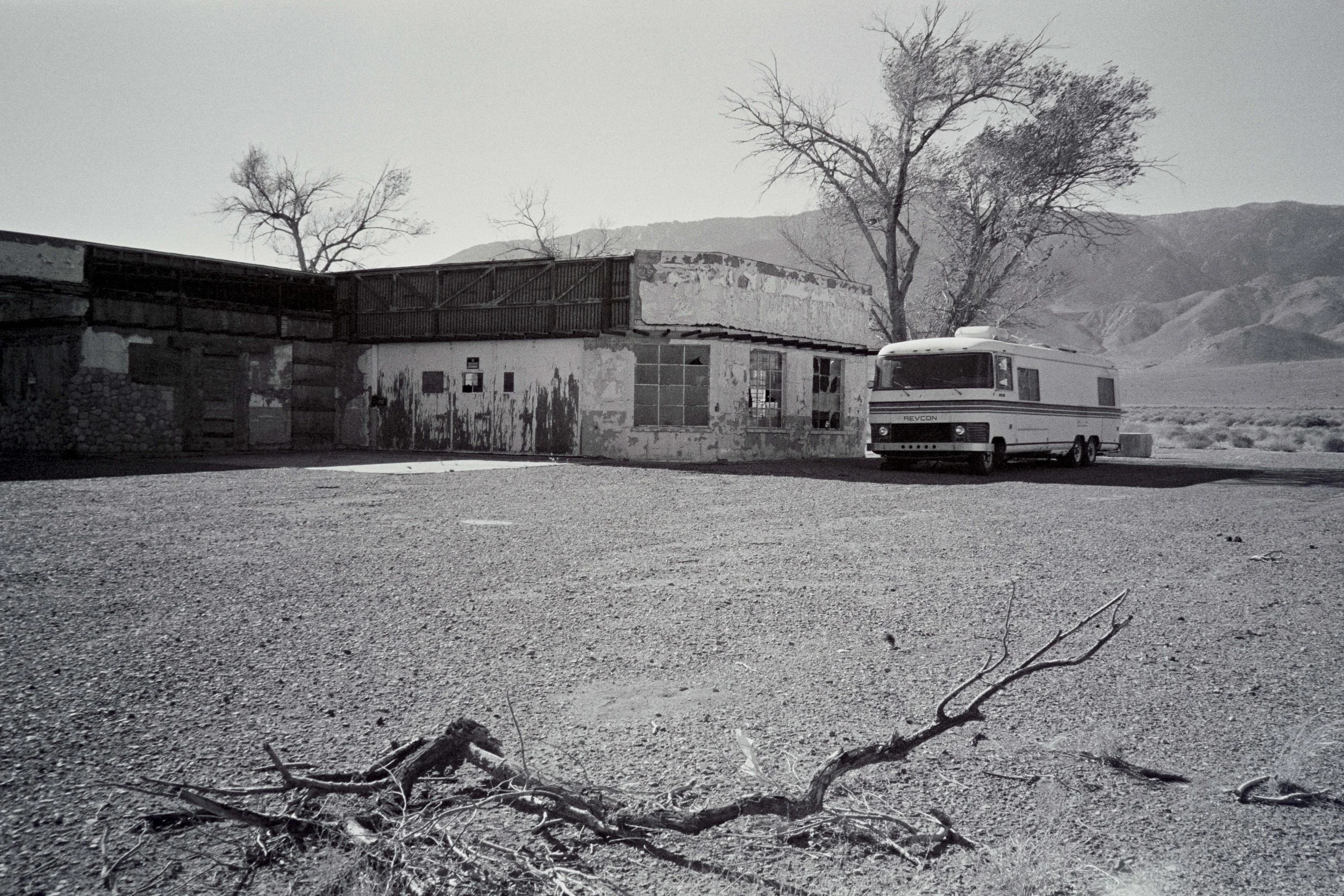Une caravane blanche stationnée devant un bâtiment abandonné dans un paysage désertique avec des montagnes en arrière-plan.