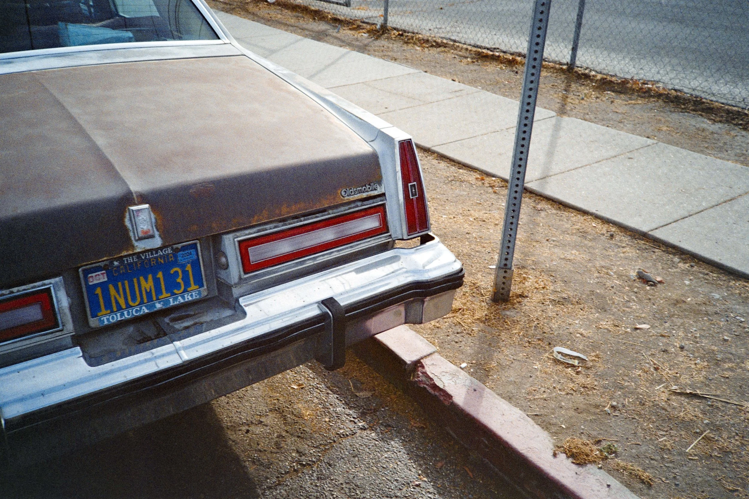 Une voiture ancienne en stationnement sur le bord de la route, vue arrière montrant une plaque d'immatriculation californiaise, une section du pare-chocs et la roue arrière, à côté d'un piquet d'arrêt de bus et d'un trottoir en béton.