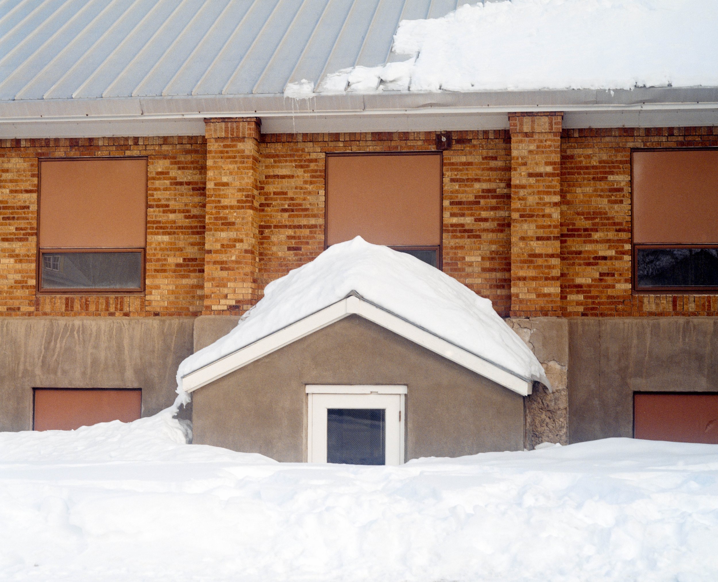 Une maison avec une façade en briques, plusieurs fenêtres avec des volets, et beaucoup de neige accumulée sur le toit et le sol.
