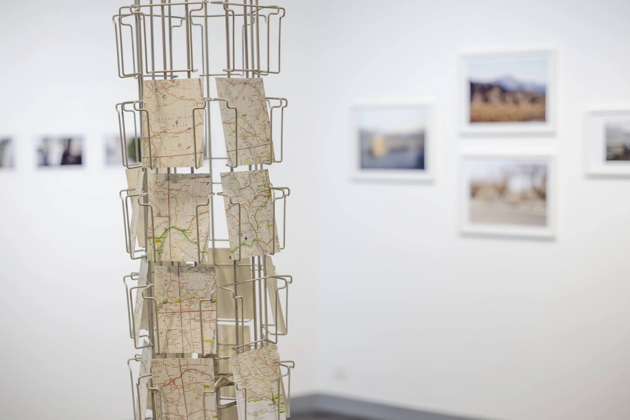 Un présentoir à postcards avec des cartes routières, dans une galerie d'art dont les murs sont ornés de photos encadrées.
