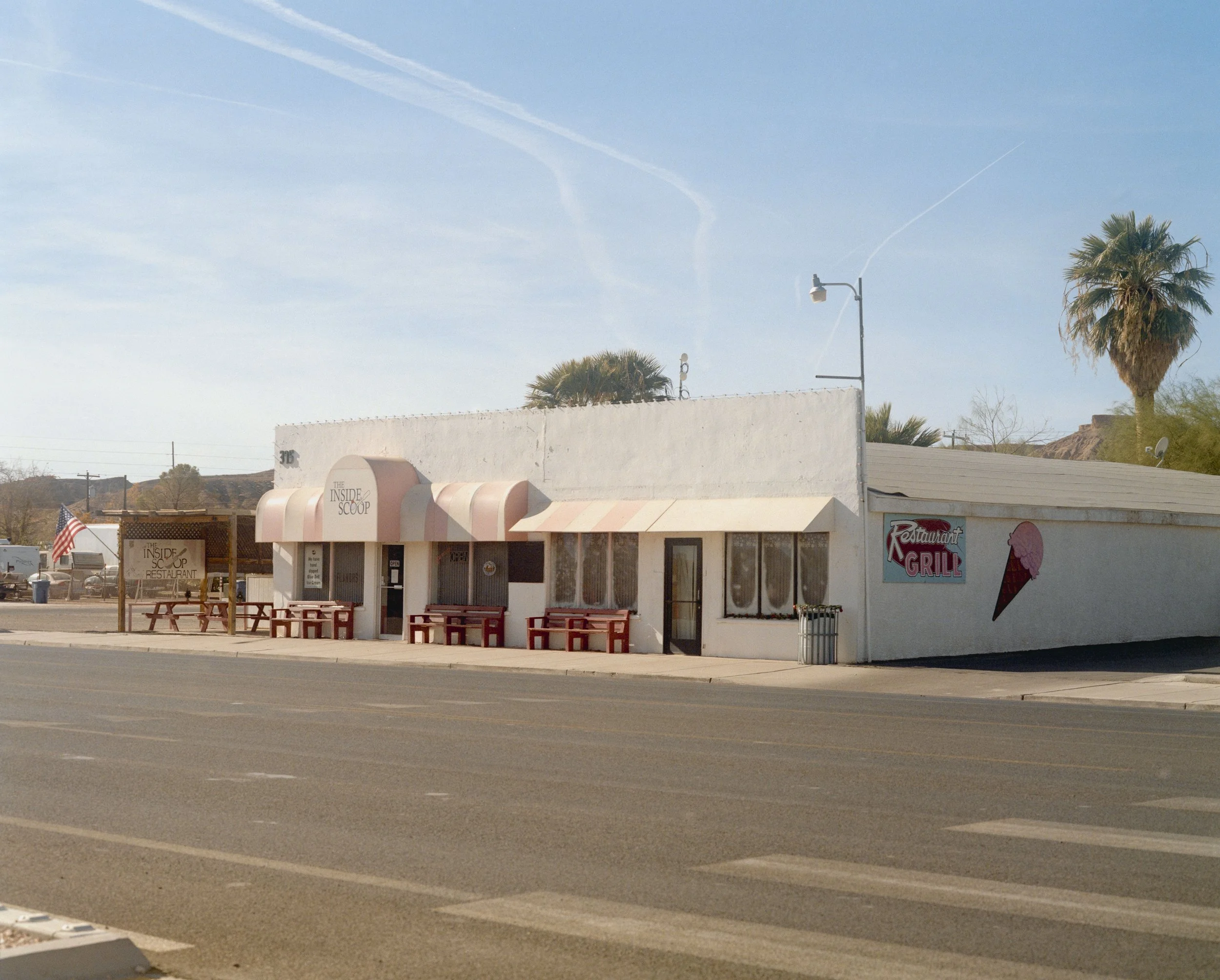 Un bâtiment blanc avec une enseigne 'The Inside Scoop' et une autre indiquant 'Restaurant Grill', des tables extérieures avec bancs rouges, un panneau en bois, des palmiers, un lampadaire, et un ciel clair avec quelques nuages.
