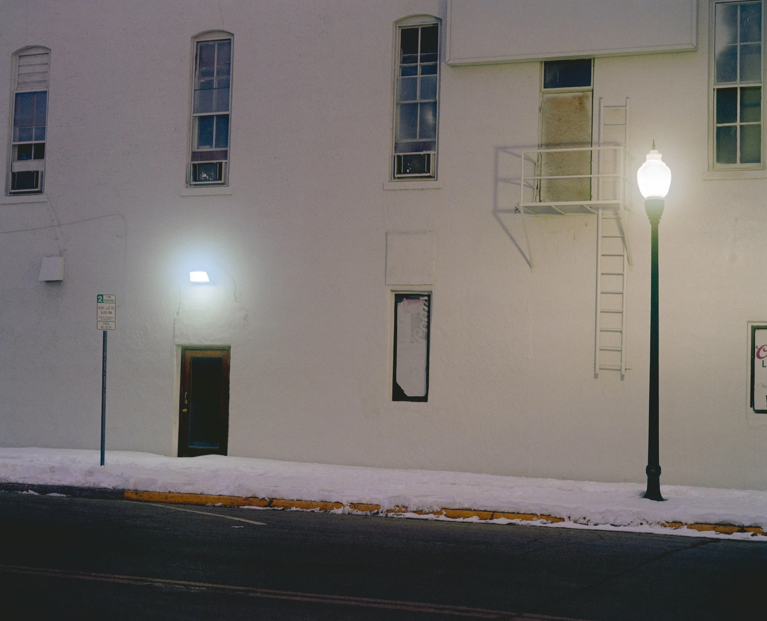 Mur blanc avec fenêtres, porte, lampadaire allumé et zone enneigée au sol.