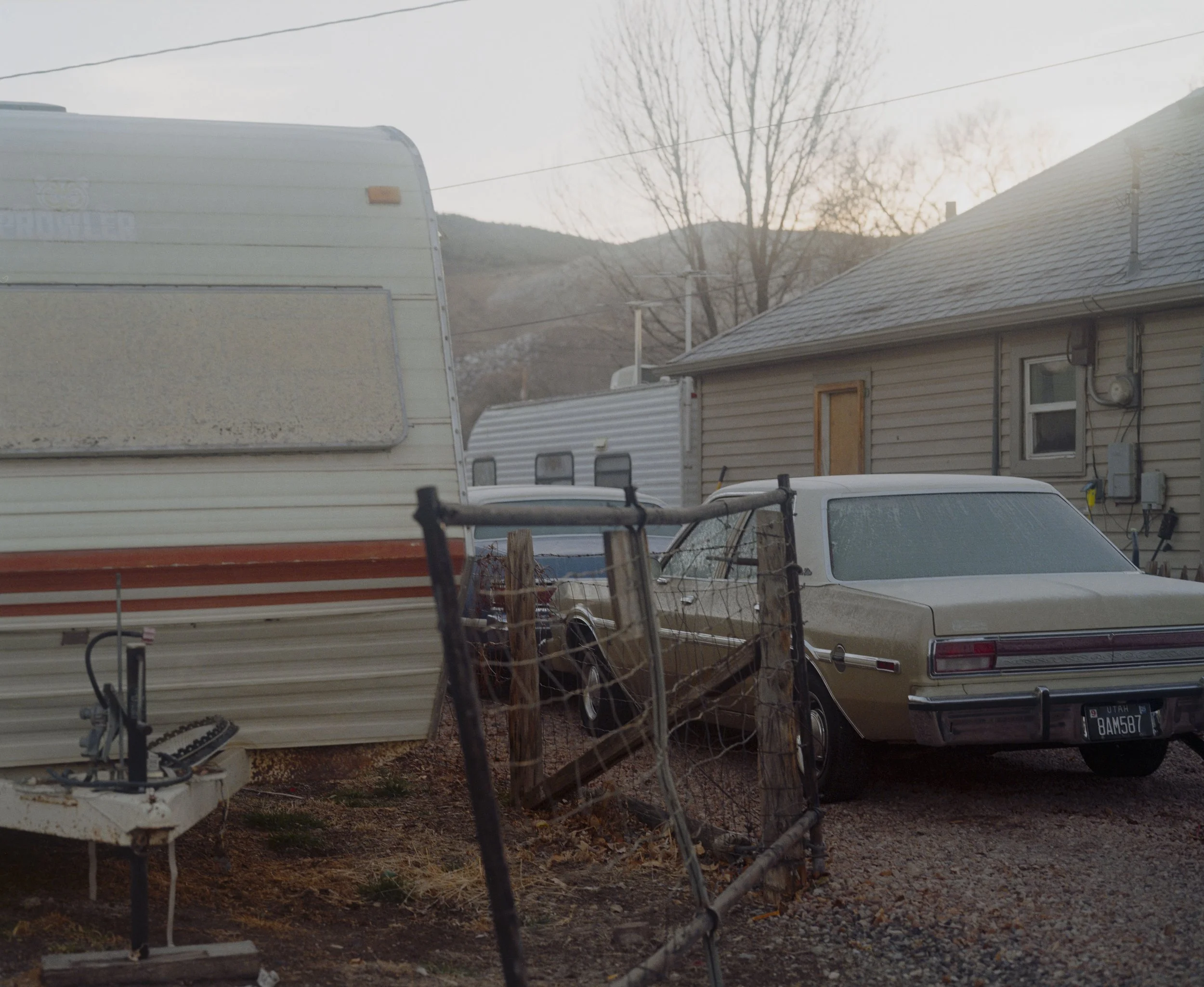 Voiture ancienne beige stationnée devant une maison avec un portail en fer rouillé, à côté d'une caravane, sous un arbre sans feuilles, avec un fond de montagnes et un ciel nuageux.
