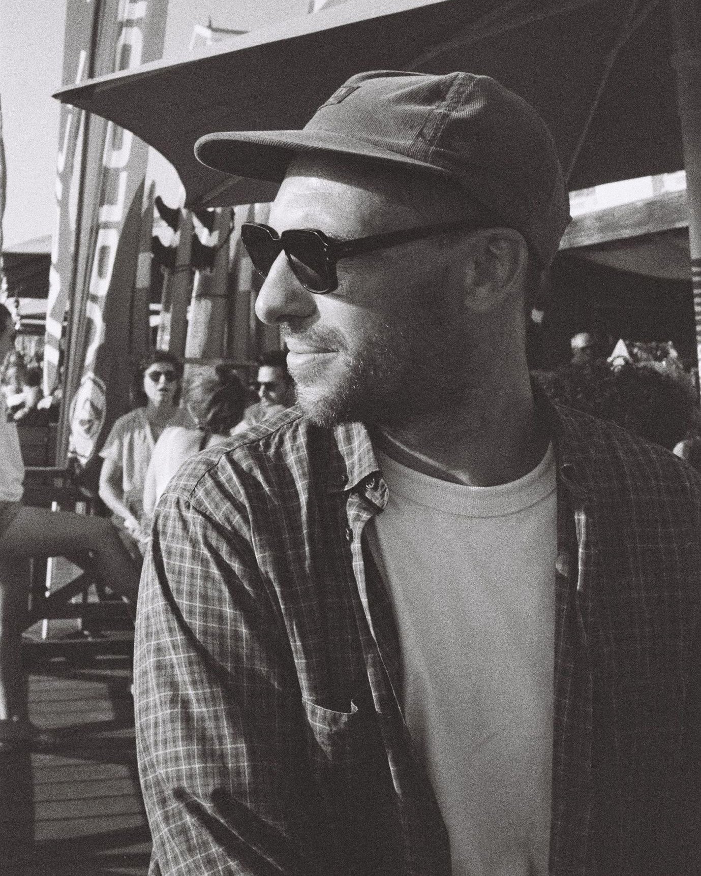 Un homme portant un chapeau et des lunettes de soleil, assis à l'extérieur avec des personnes en arrière-plan