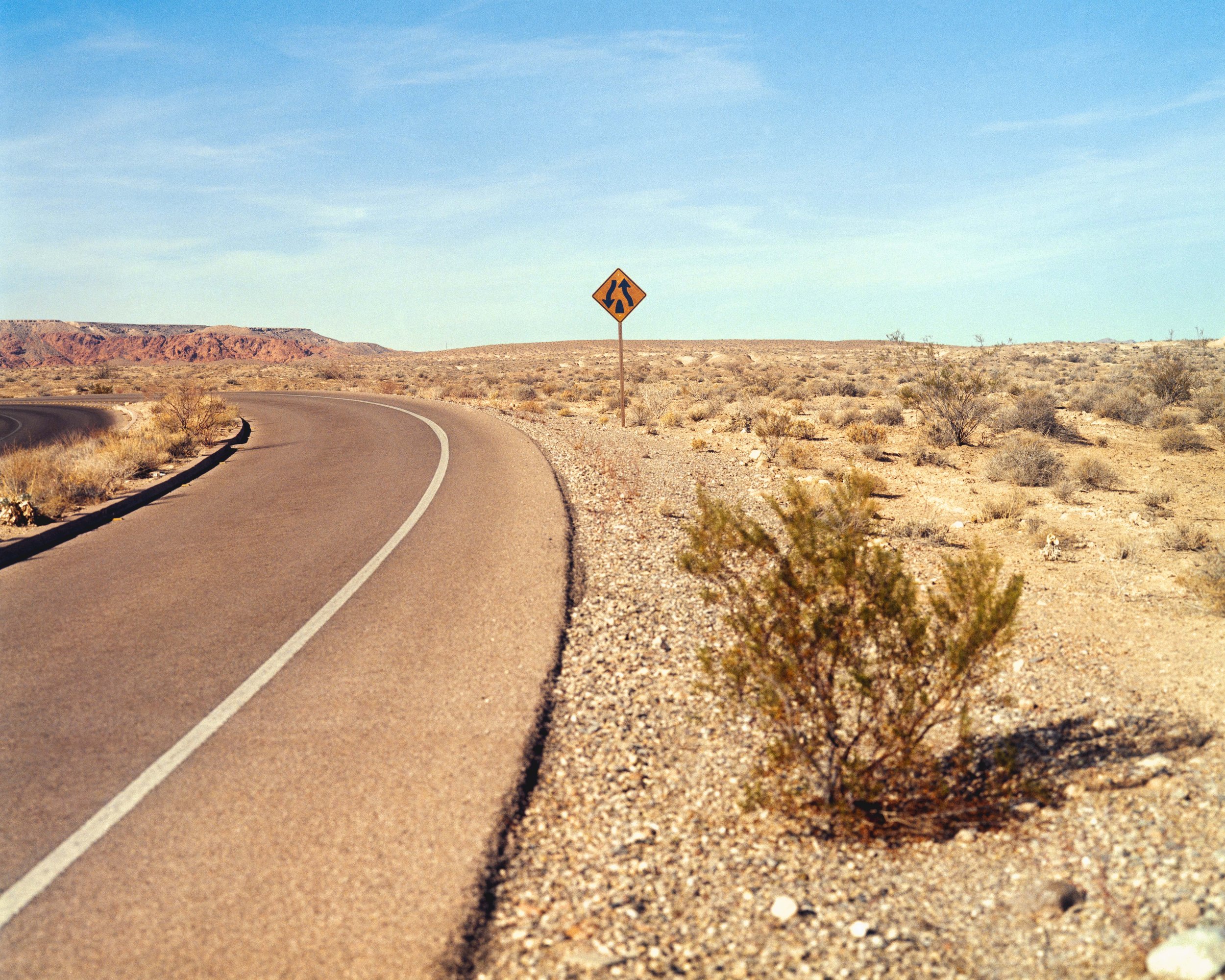 Route désertique avec panneau de signalisation sur le côté, terrain aride avec petits arbustes, ciel bleu clair.