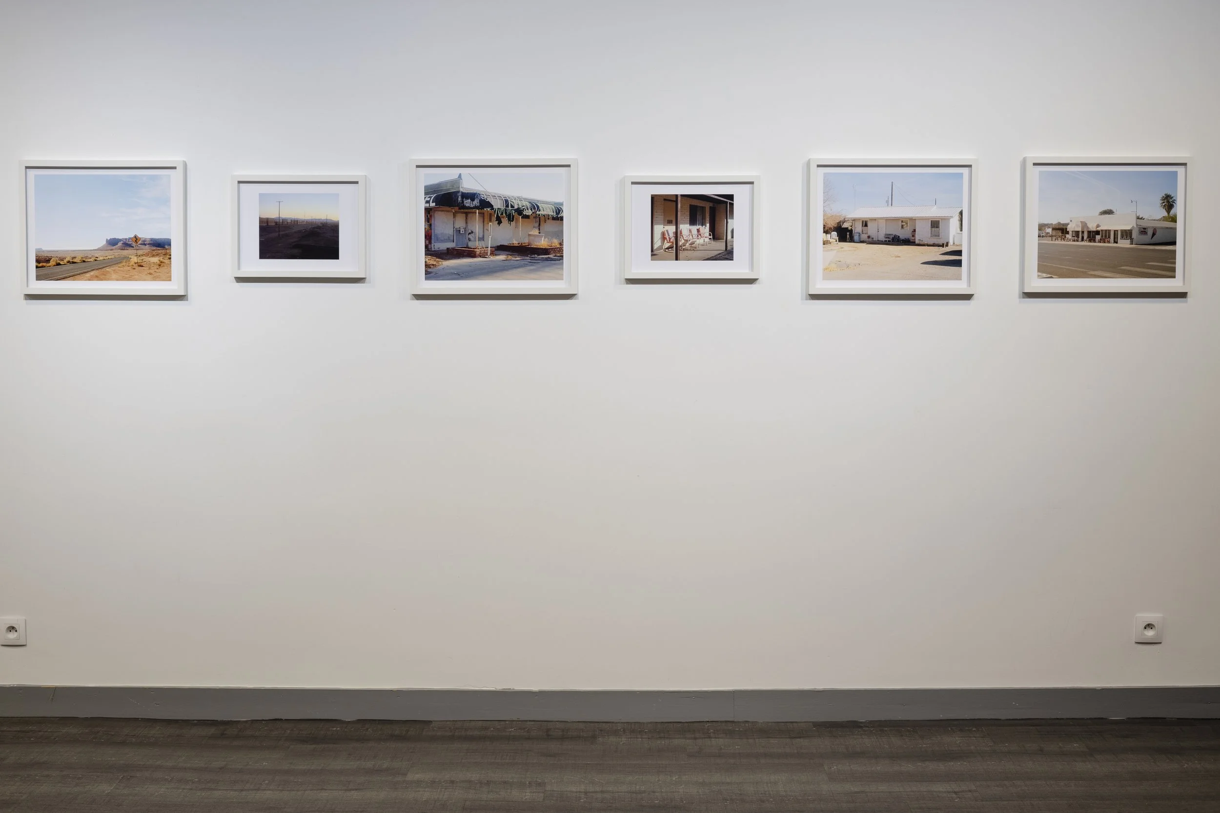 Une série de sept photographies encadrées accrochées à un mur blanc, représentant des scènes de paysages et de bâtiments dans un environnement désertique ou rural.