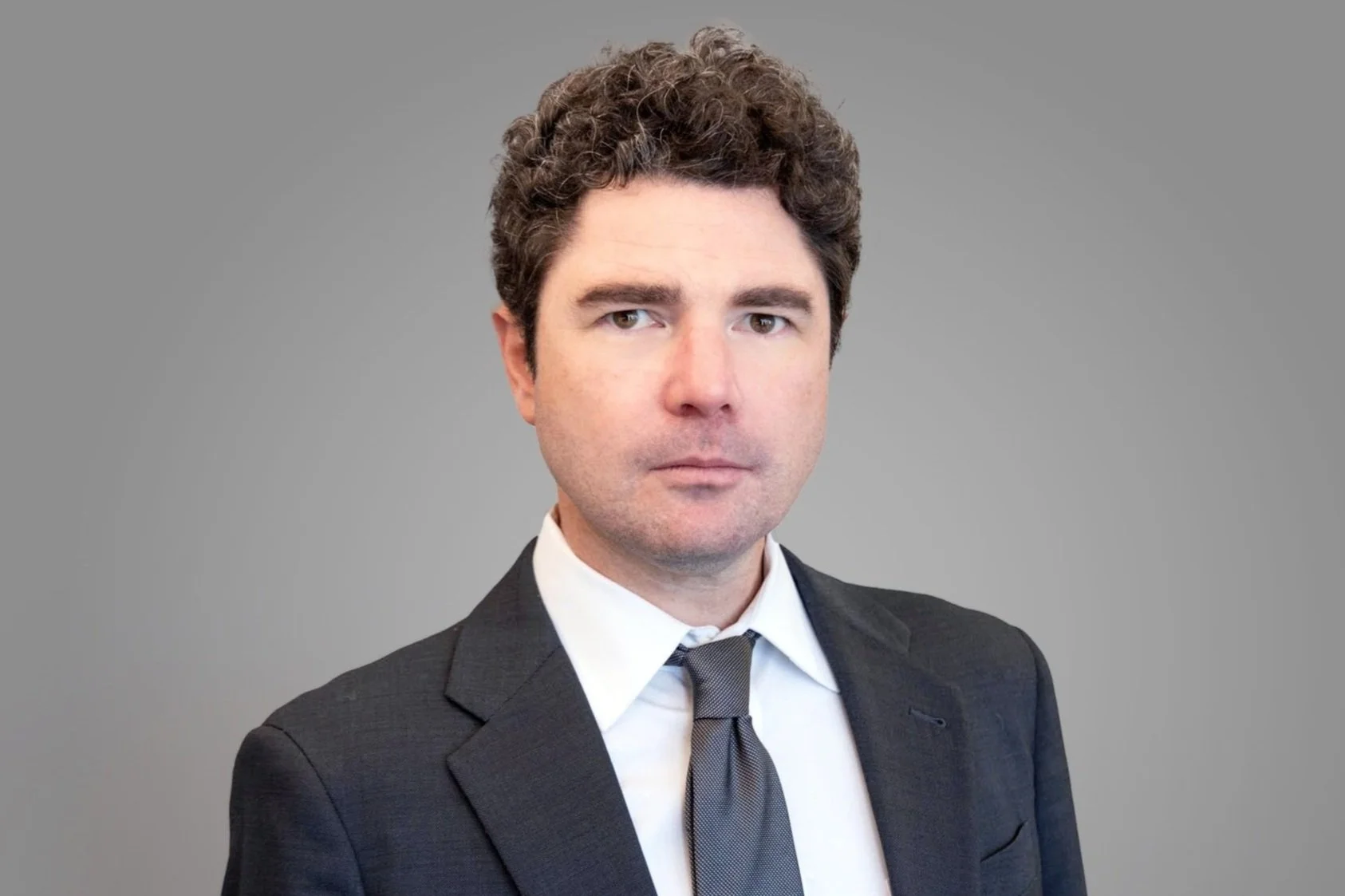 Professional portrait of a man with curly brown hair wearing a dark suit, white shirt, and a gray tie against a plain gray background.