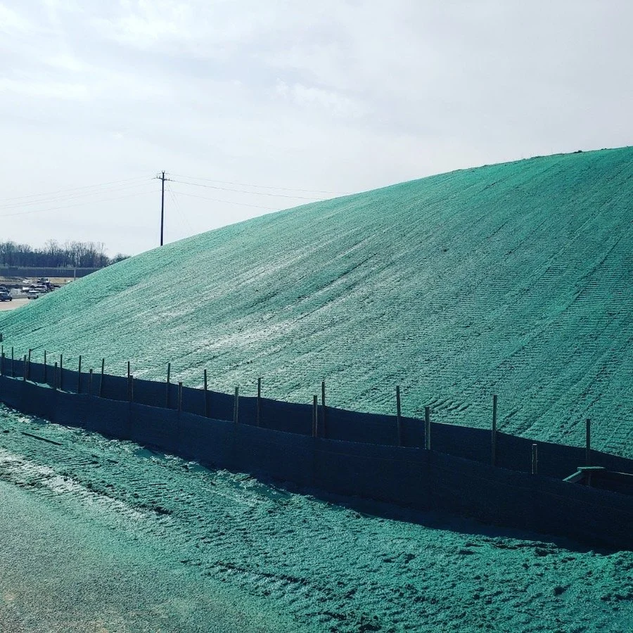 A large slope covered in green hydroseeding with a silt fence at the base, against a cloudy sky backdrop.