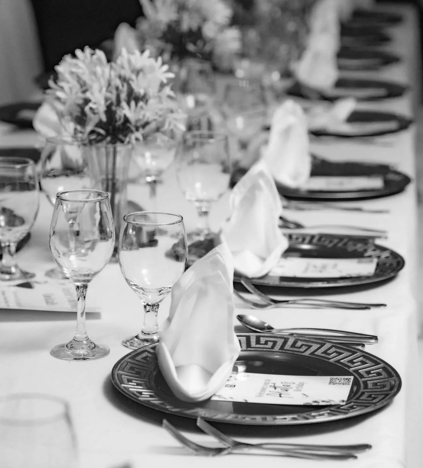 A black and white photo of a formal dining table set for a meal, featuring plates, wine glasses, folded napkins, and a floral centerpiece.