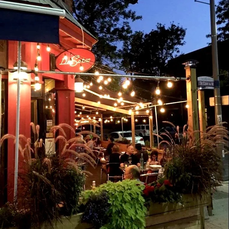 Nighttime view of Desotos Eatery's side patio on the corner of St. Clair West and Lauder Ave, featuring bright string lights, vibrant red walls, and the illuminated Desotos hanging sign.