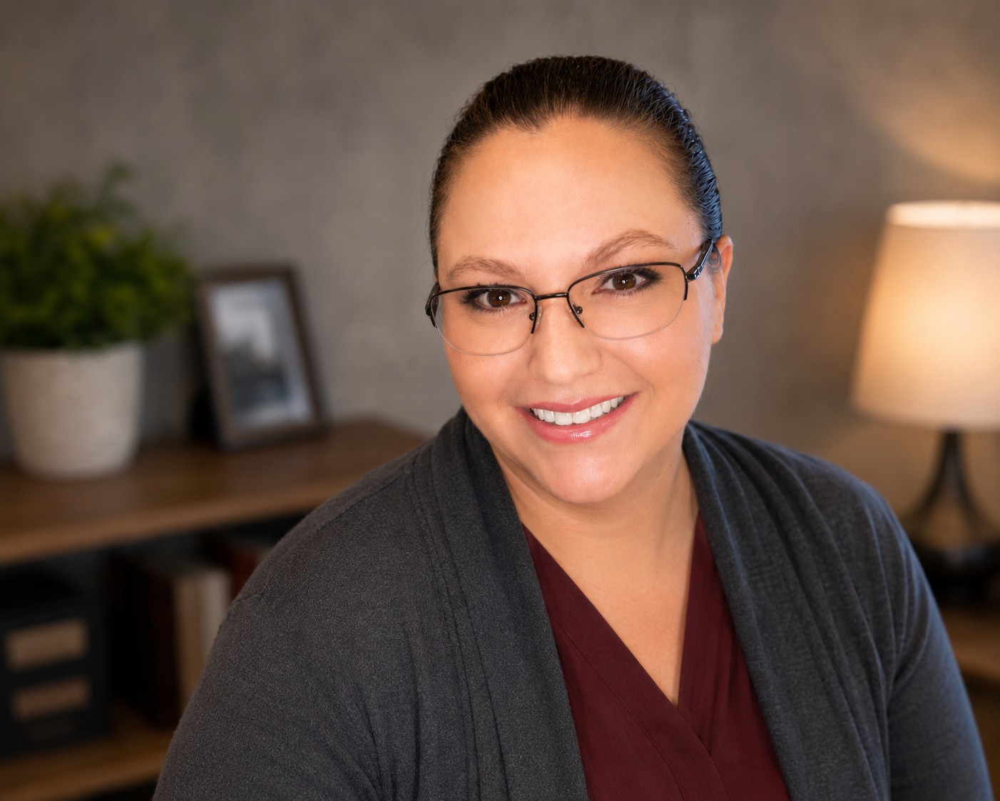 A woman with glasses and dark hair tied back, smiling, sitting in a cozy room with a lamp and framed photo in the background.