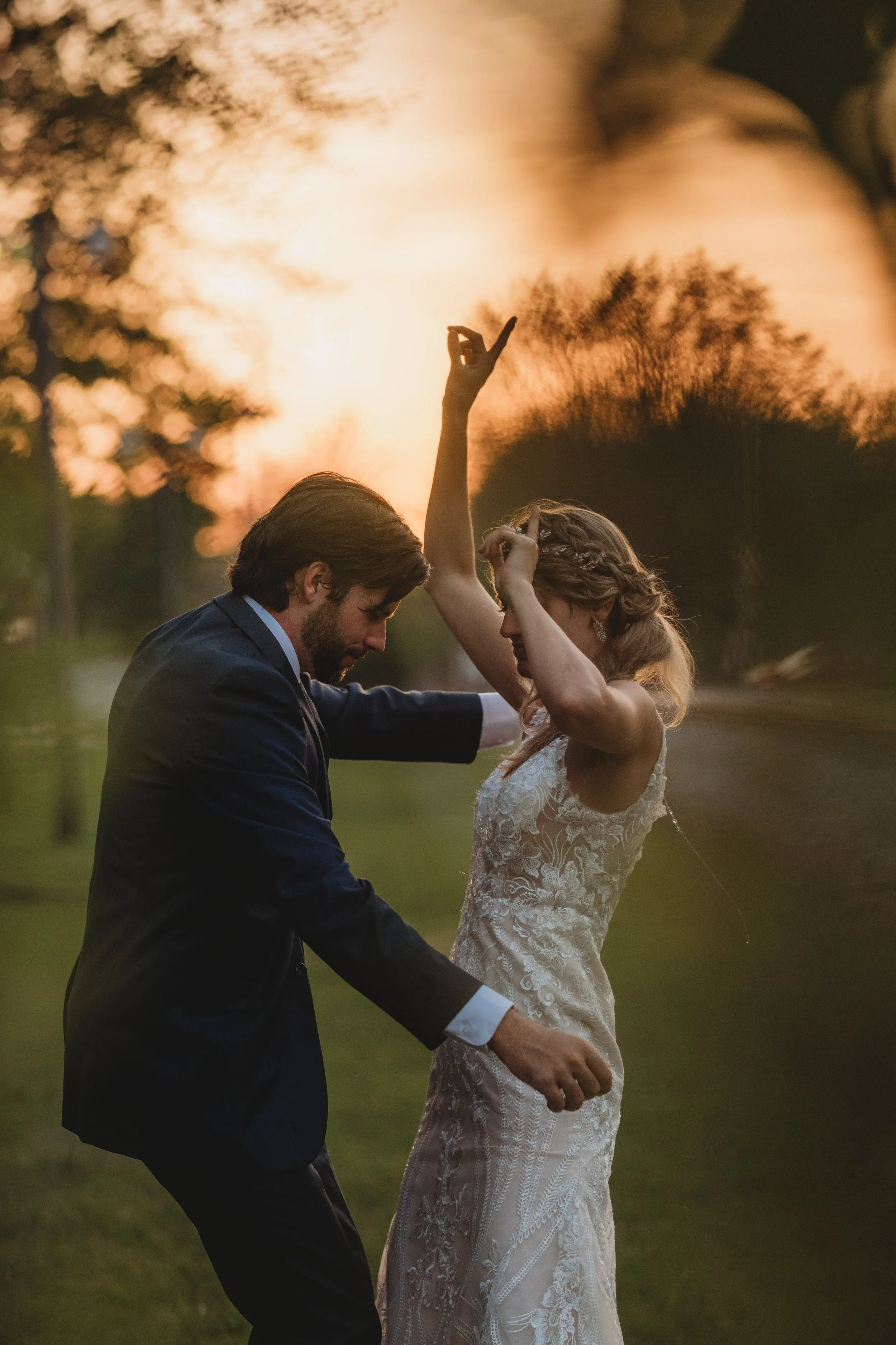 A wedding couple dancing outdoors at sunset with trees and warm sky in the background.