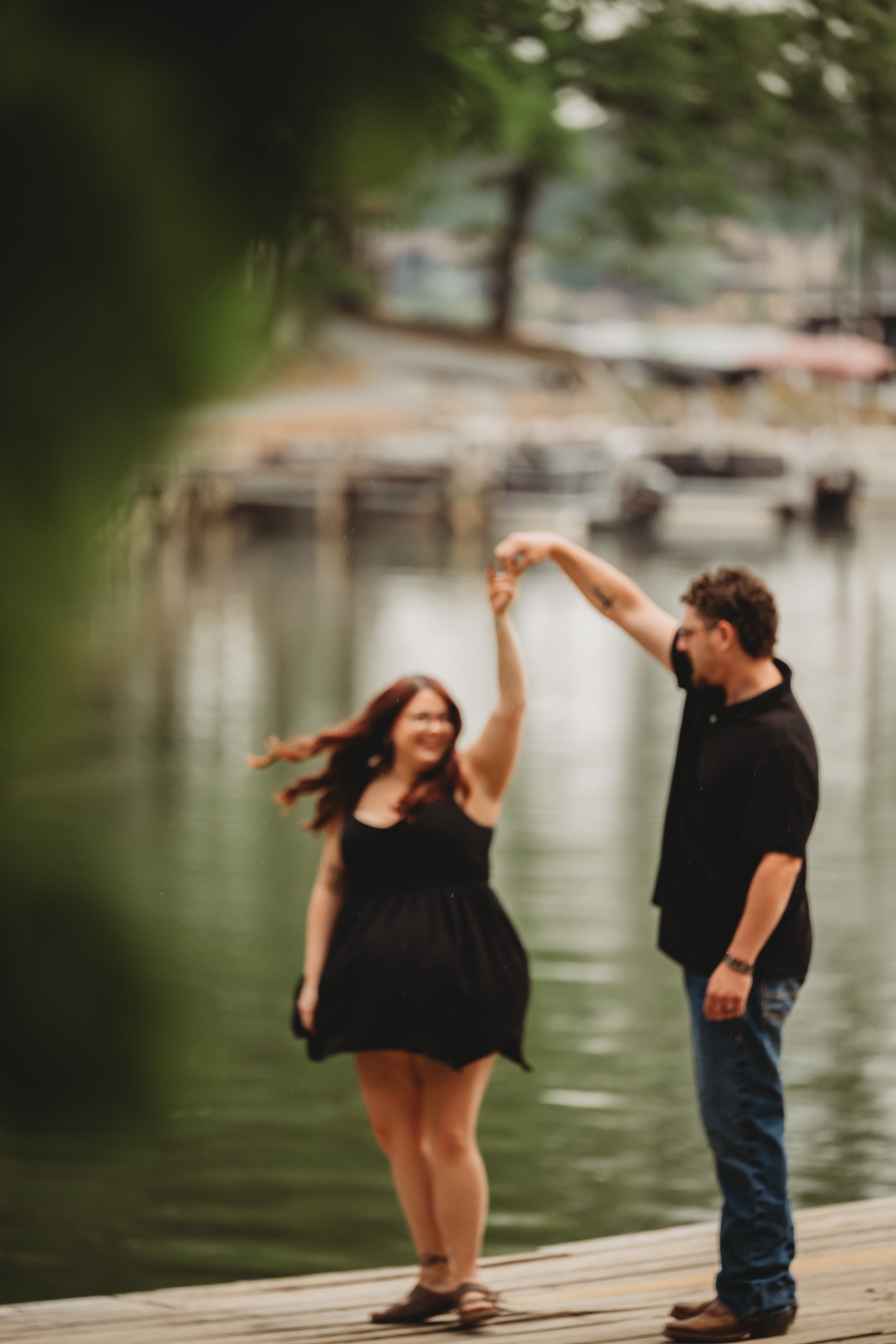A couple dancing on a wooden dock by the water, with trees and boats in the background, and their faces are not visible.