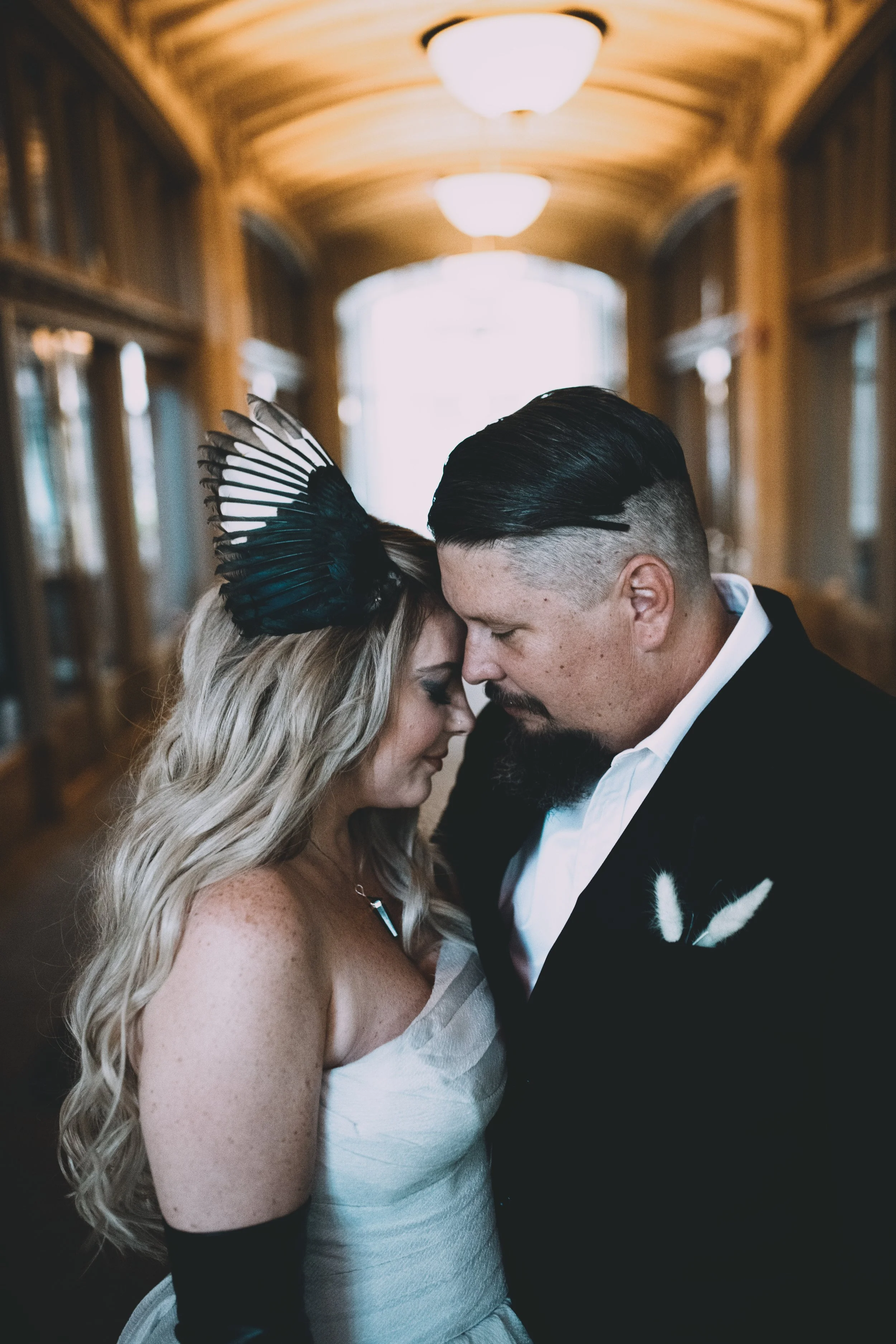 A couple dressed in wedding attire holding foreheads together in an indoor wooden corridor with warm lighting.