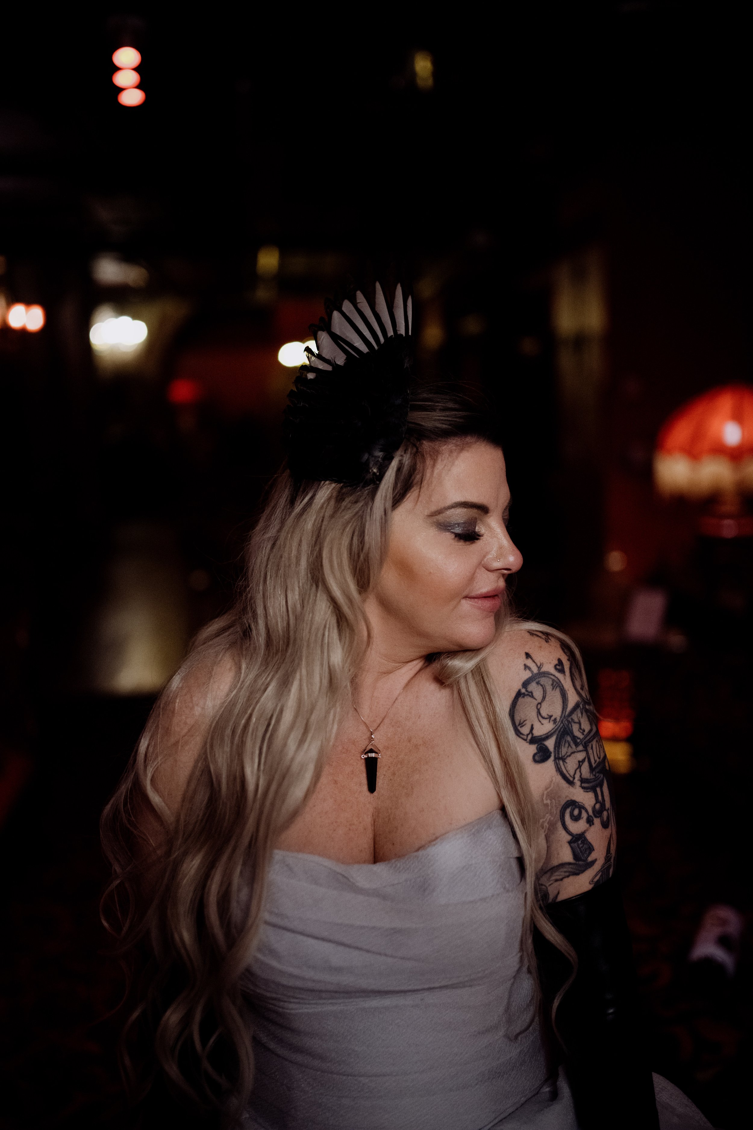 A woman with long wavy blonde hair with tattoos on her upper arm, wearing a light gray strapless top, silver necklace, and black feather headband, sitting indoors at night with blurred colorful lights in the background.