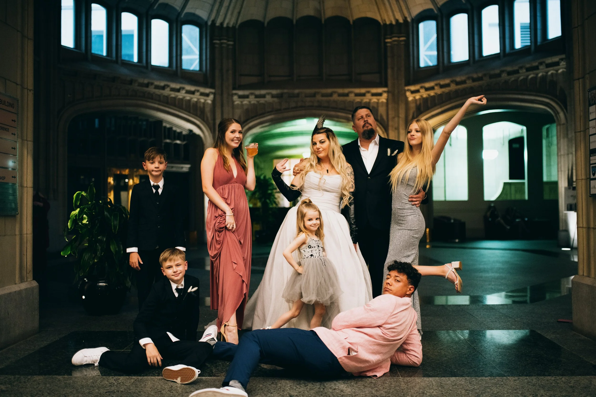A group of people, including children and adults, posing in an indoor setting with an arched ceiling and large windows. Some are standing, while others are lying or sitting on the floor, showcasing a casual, playful atmosphere.