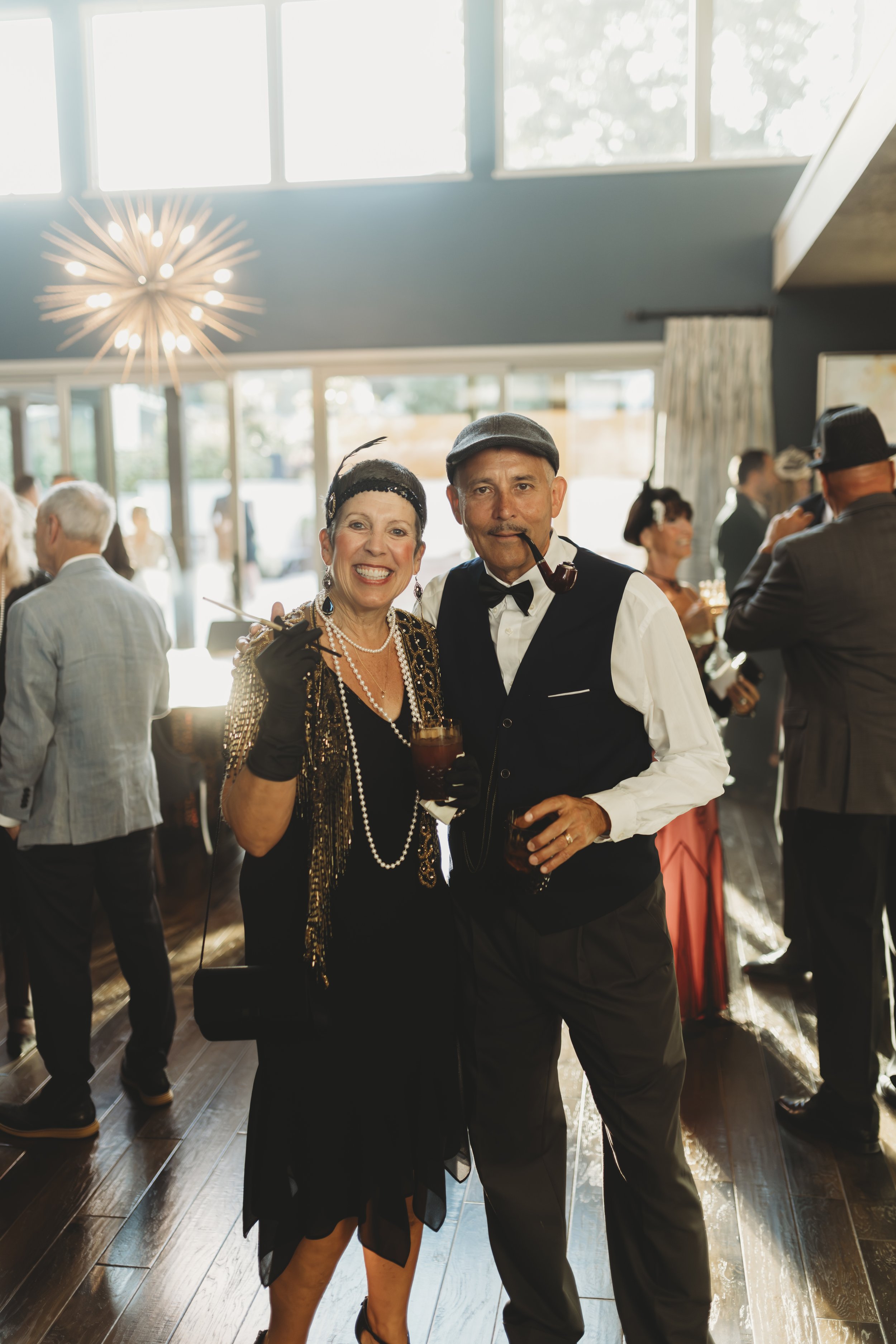 Two people at a social gathering dressed in vintage or 1920s style clothing, smiling and holding drinks, with other guests in the background and natural light coming through large windows.