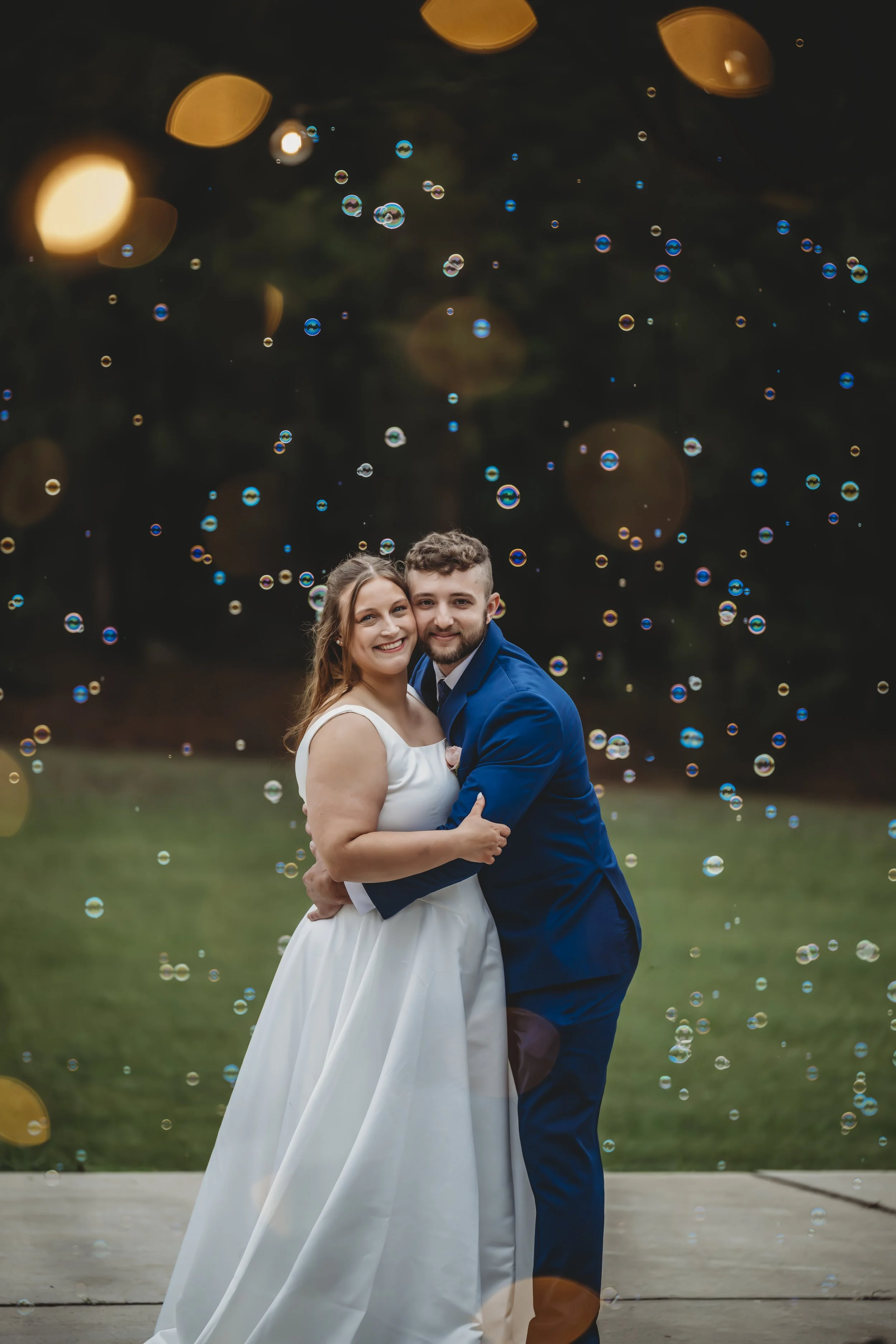 A smiling bride and groom embracing outdoors at sunset, surrounded by floating soap bubbles with blurred lights in the background.